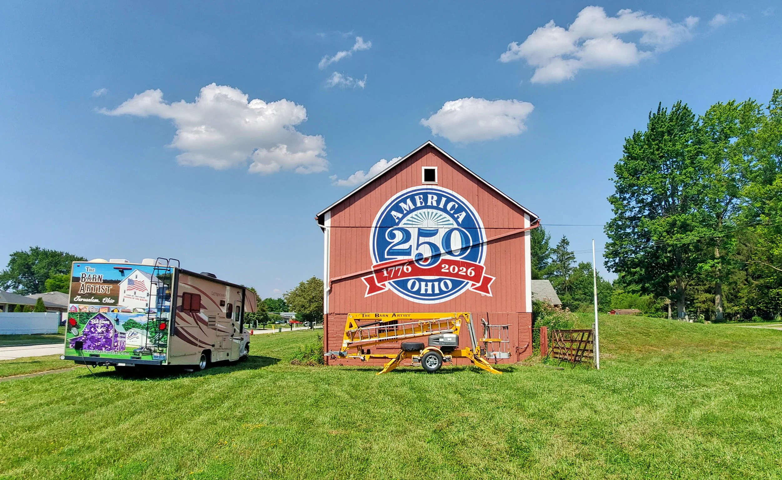 A red barn celebrating America's 250th anniversary with a large sign that reads "America 250 Ohio 1776-2026." In front of the barn, there are various vehicles and equipment, including a colorful RV and construction tools, with green grass and trees u