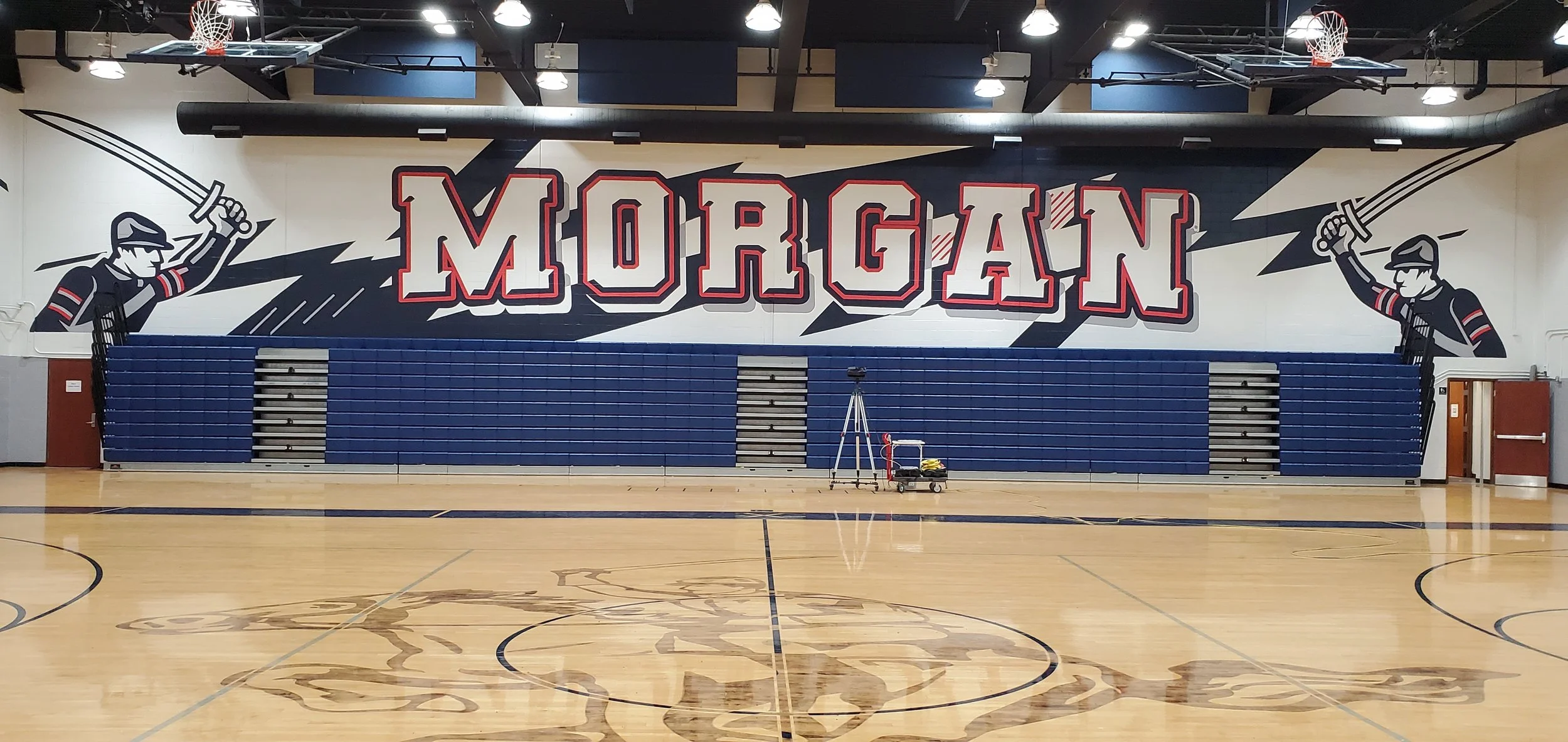 A gymnasium with a large wall mural featuring the word 'MORGAN' in bold red and white letters, flanked by illustrations of a male figure holding a sword on each side, with sectioned blue gym bleachers below the mural and a hardwood floor with a decor
