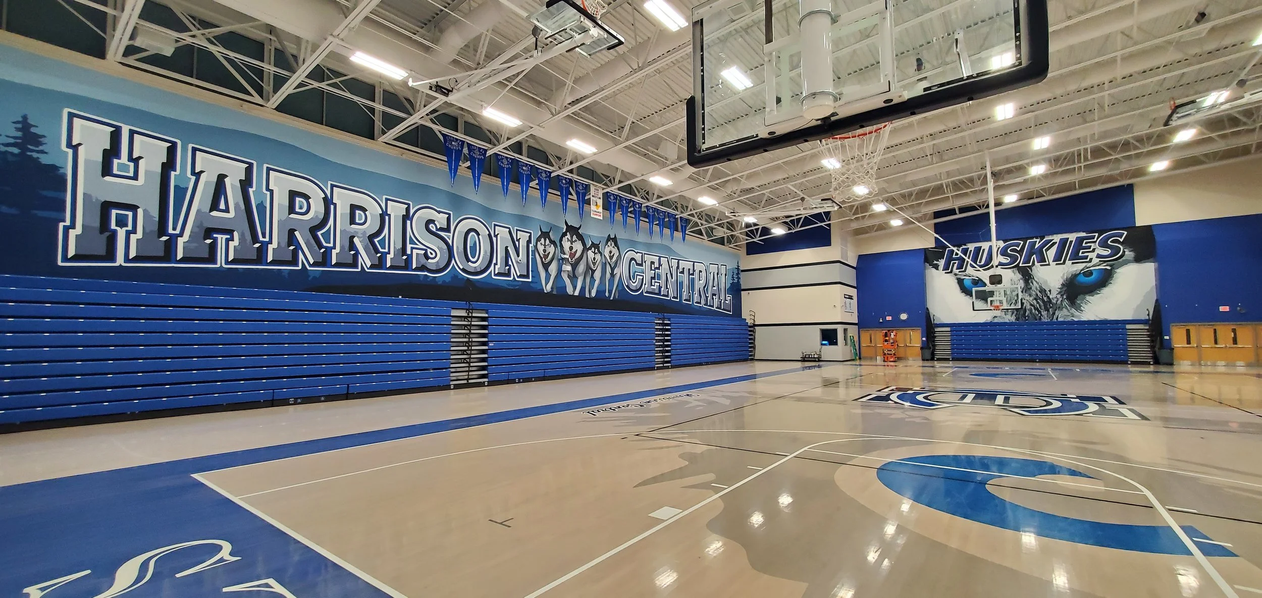 Empty indoor school gymnasium with blue bleachers, basketball hoop, and large murals of huskies and the words 'Harrison Central' on the walls.