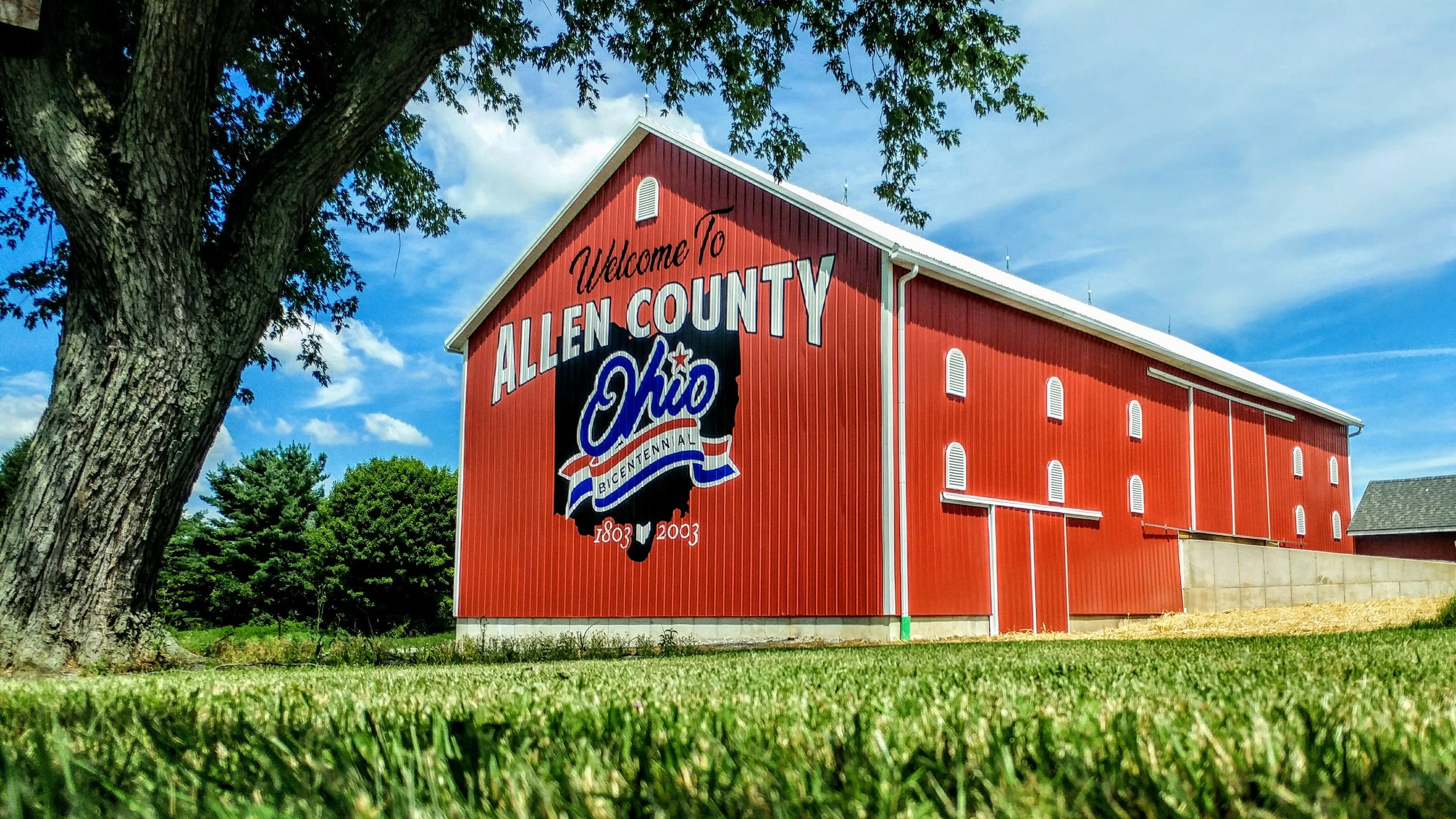 A red barn with a mural that says "Welcome To Allen County, Ohio, Bicentennial 1803-2003" on the side, situated on a grassy area with trees and a blue sky with clouds.