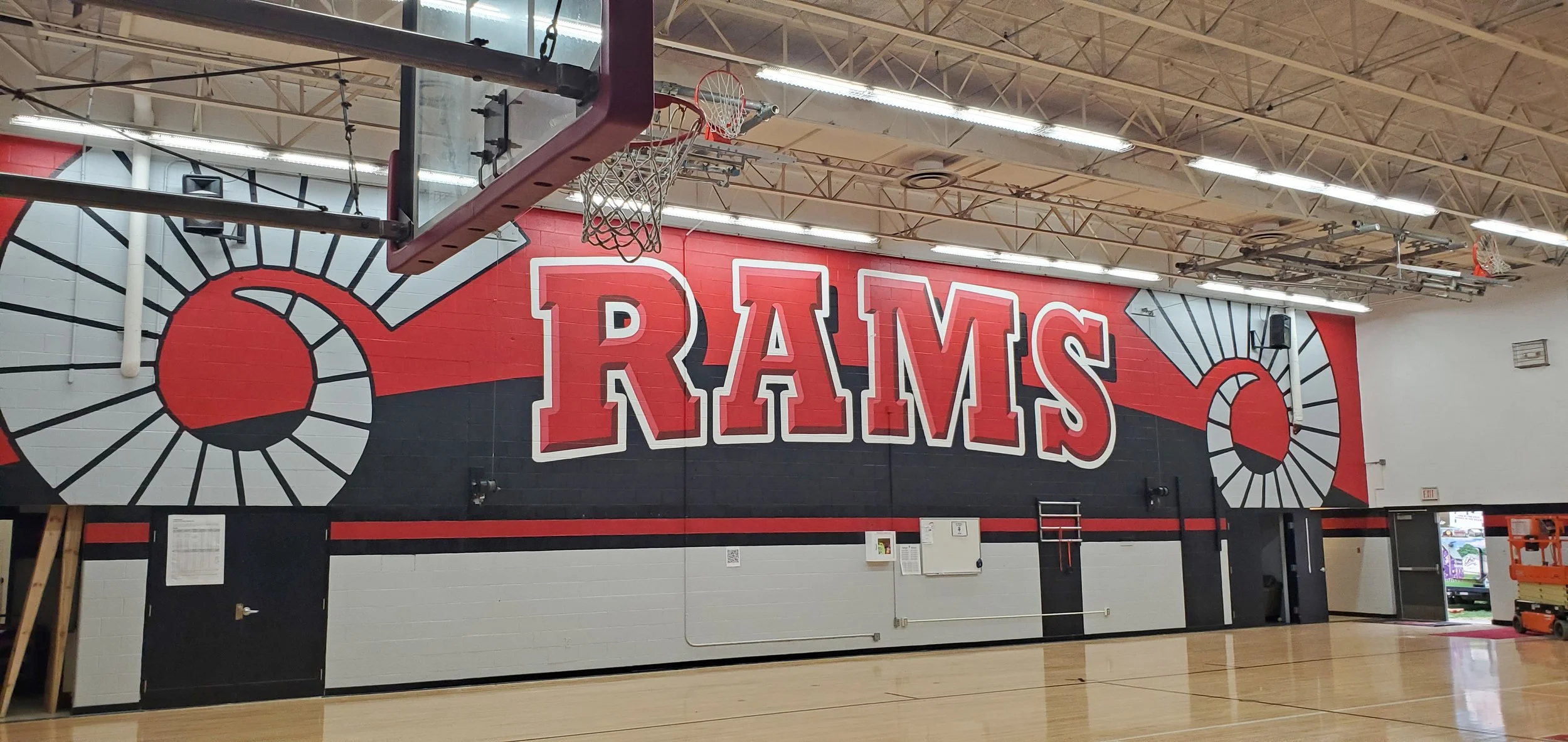 An indoor basketball court with a mural of the Los Angeles Rams logo on the wall. The mural features the word 'RAMS' in red letters, with images of football helmets on each side.