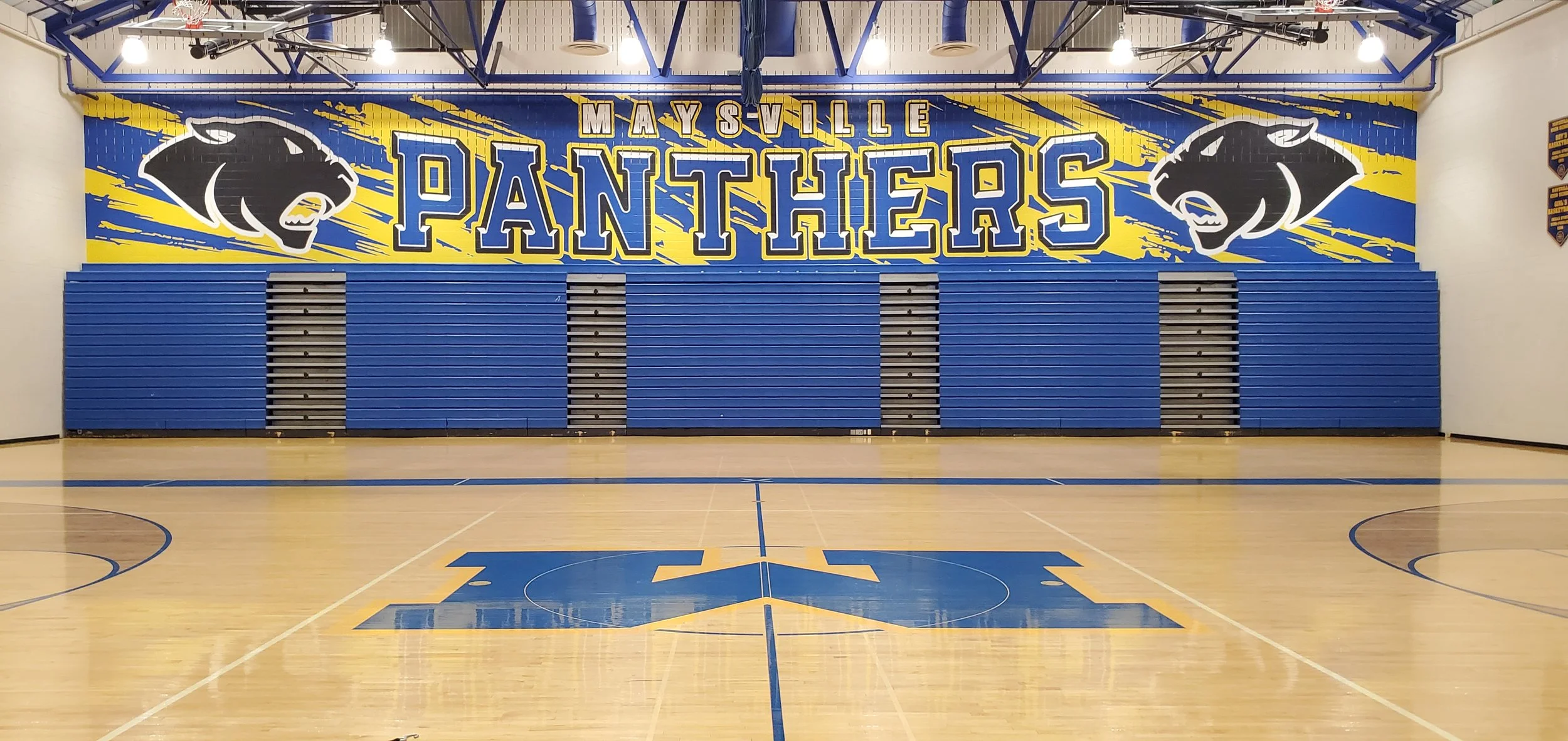 Empty basketball gymnasium with closed blue bleachers and a large mural wall featuring the words 'Mayville Panthers' with panther mascots on each side in black and white. The gym has polished wood floors, basketball court markings, and bright overhea