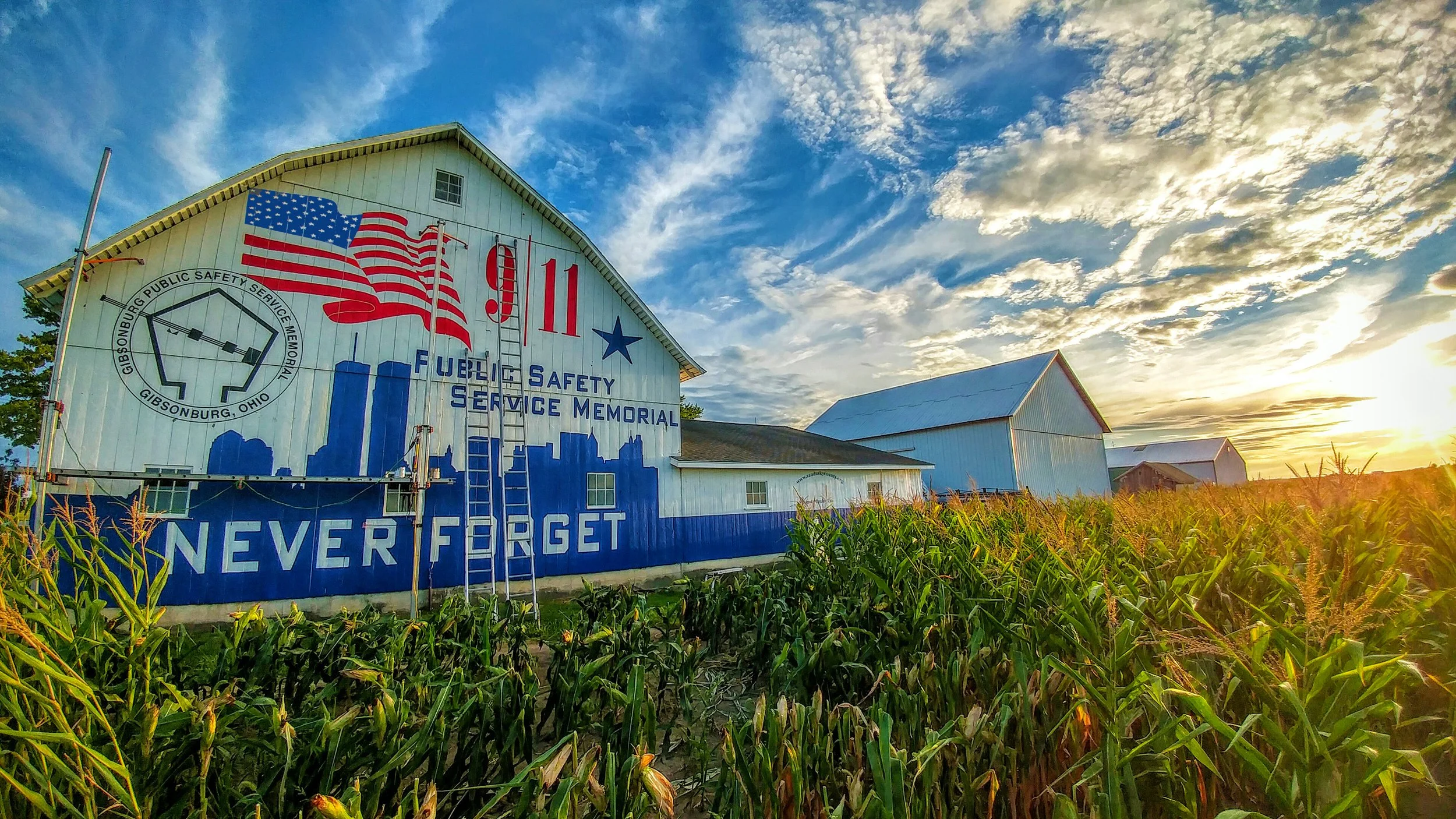 A barn with patriotic decorations and the words "Never Forget" painted on it, set in a field of corn at sunset.
