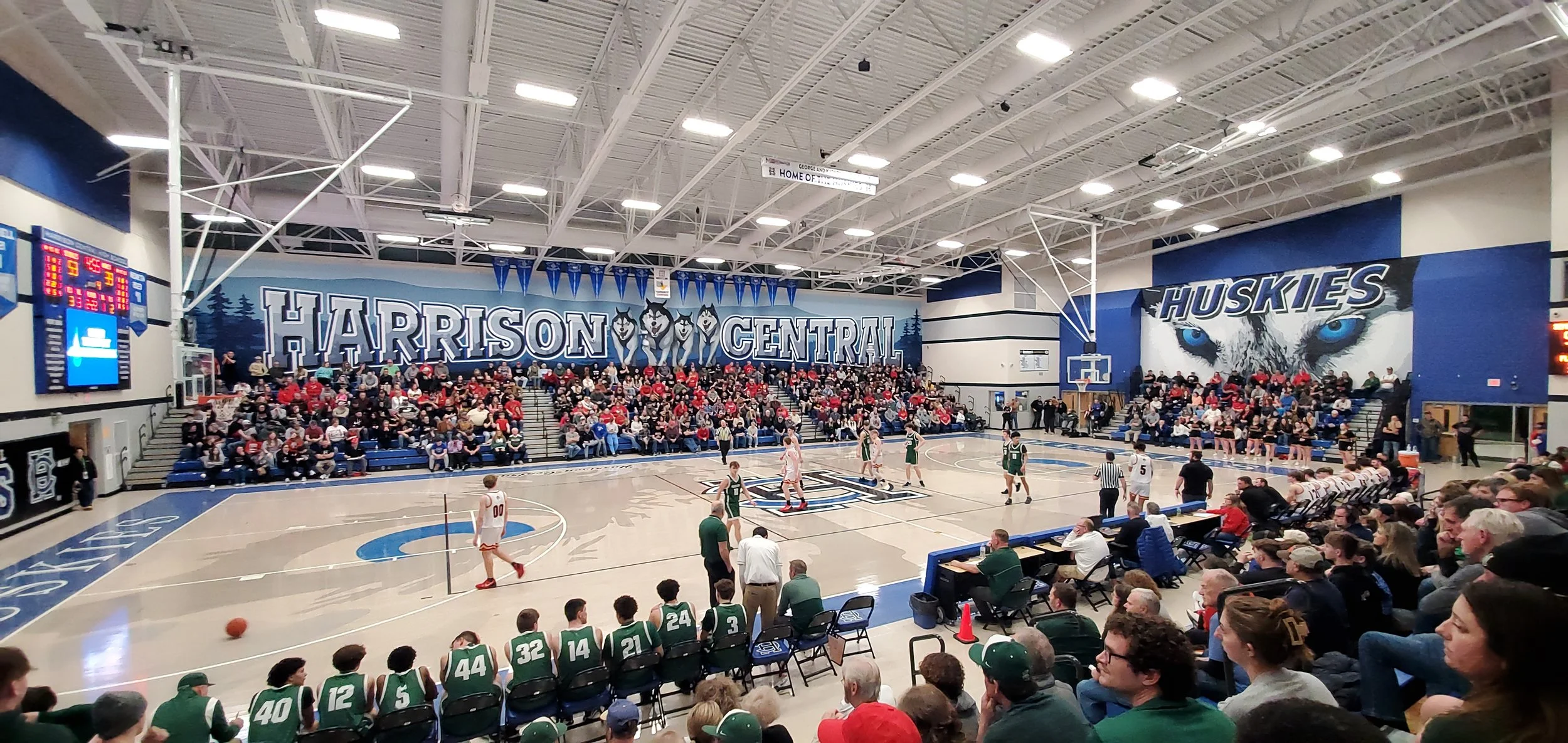 Basketball game inside Harrison Central gymnasium with players on the court, cheerleaders, and many spectators in the stands. Large wall murals feature wolf mascots and the words 'Harrison Central' and 'Huskies'.