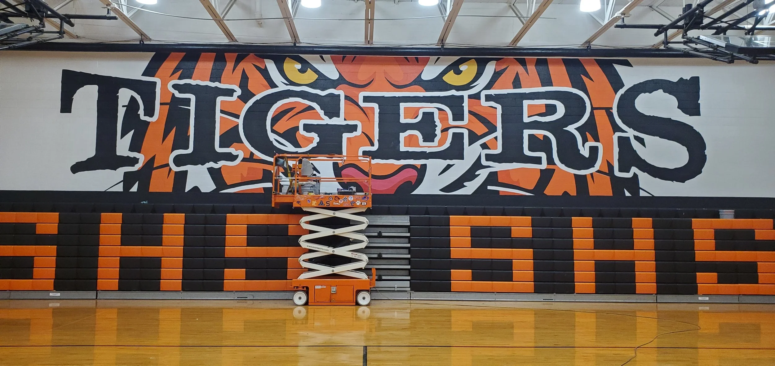 A gymnasium wall painted with a large tiger face behind the word 'TIGERS' in bold letters, with a person on a scissor lift in front of the wall.