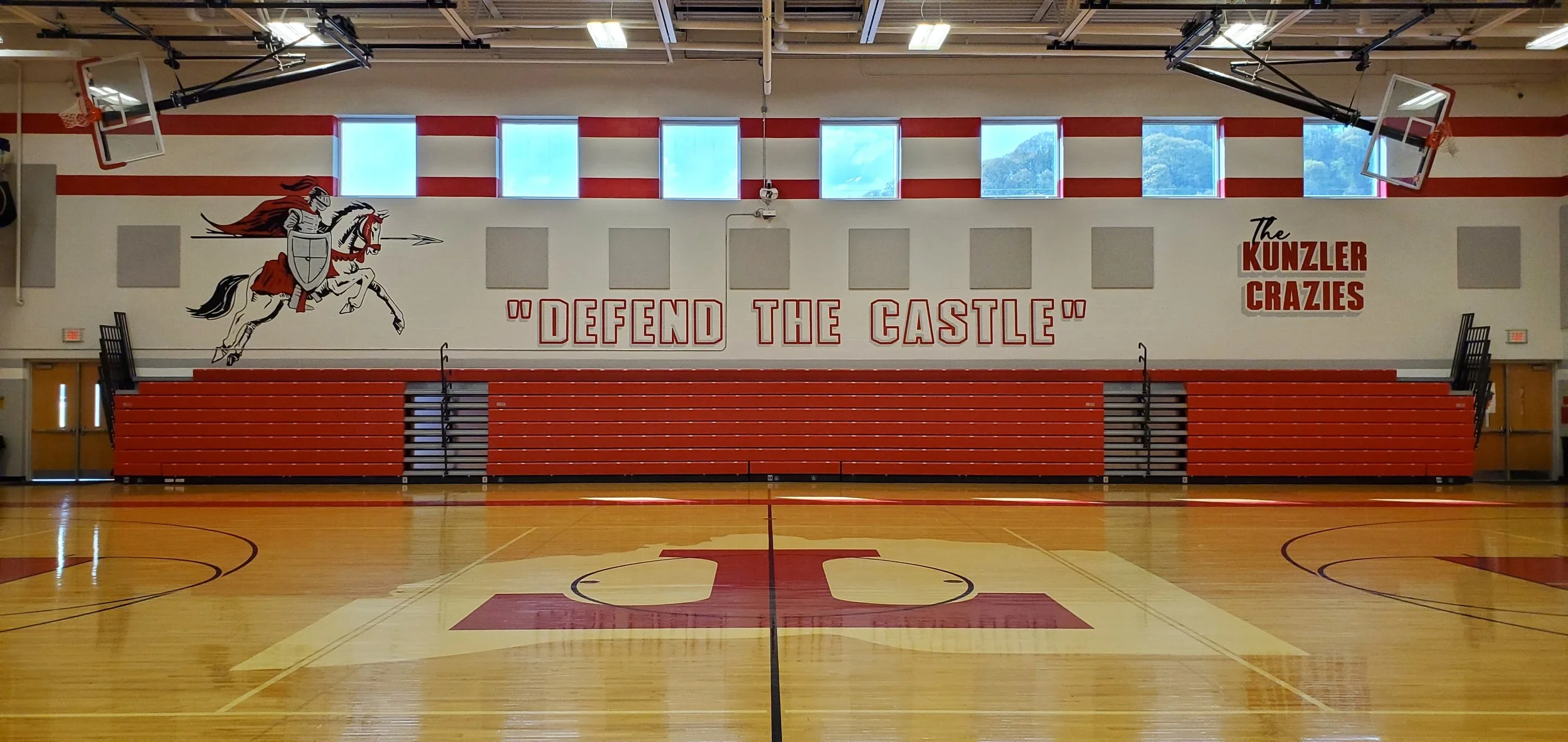 A gymnasium with a basketball court, red bleachers, and a large wall featuring a graphic of a knight on a horse and the words 'Defend the Castle' and 'The Kunzlzer Crazies'.