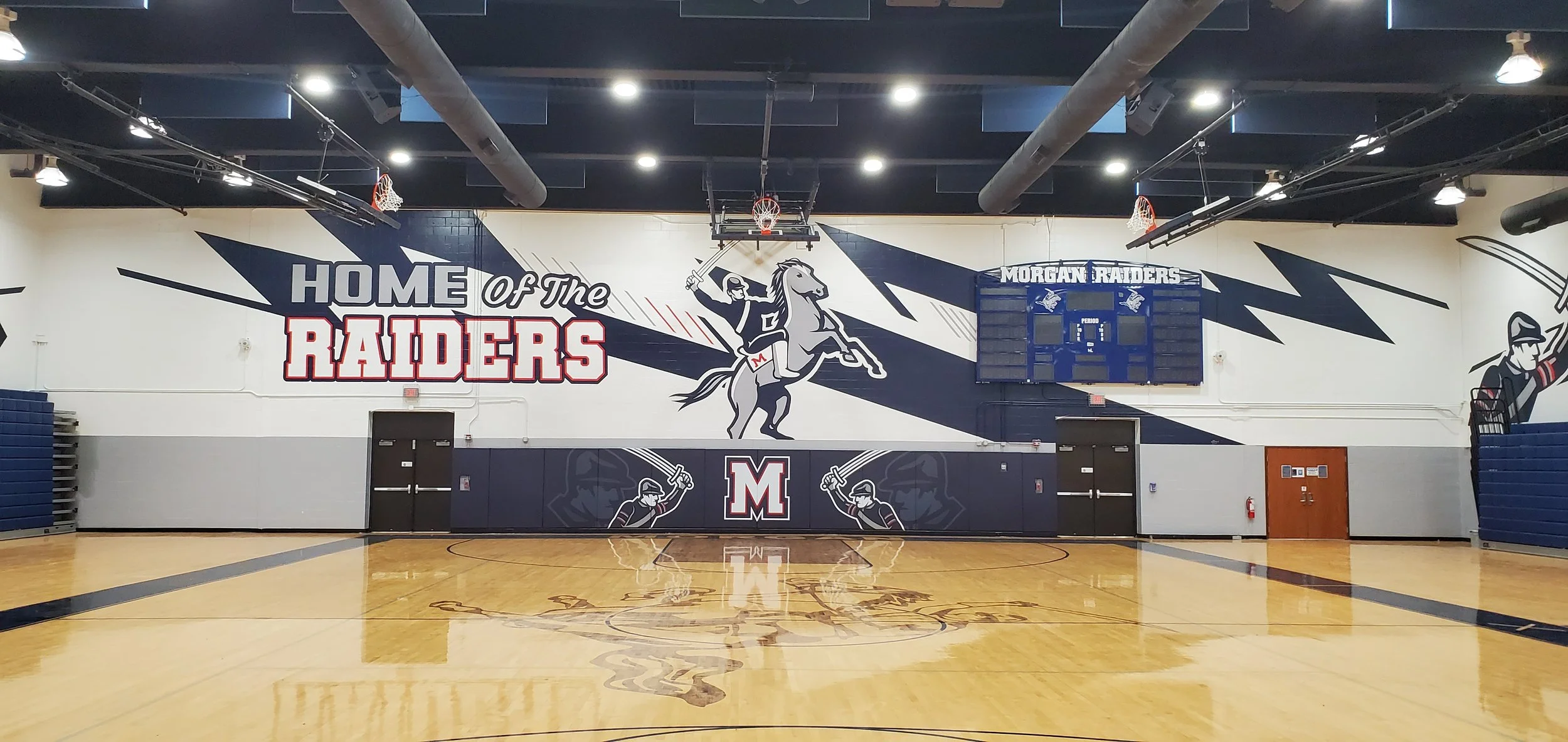 Interior of a gymnasium with a large mural on the wall that says 'Home of the Raiders' and features a cartoon knight riding a horse, holding a sword, with a scoreboard labeled 'Morgan Raiders' above it. The gym has blue basketball hoops, a polished w