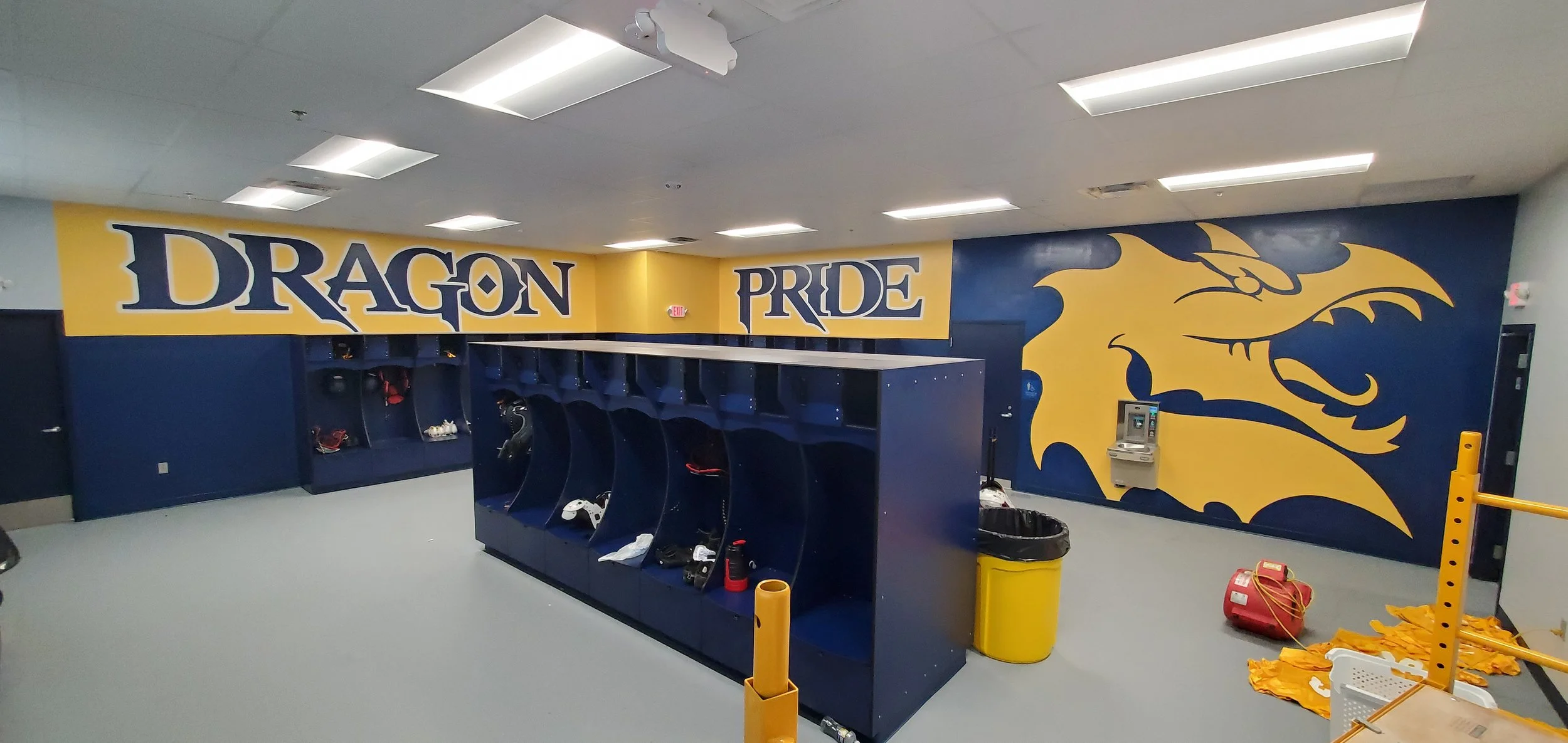 A locker room with a mural on the wall reading 'Dragon Pride' and a dragon illustration on the right side. The room has blue lockers, a water fountain, and a few scattered items such as a trash can, a red fire extinguisher, and cleaning supplies.