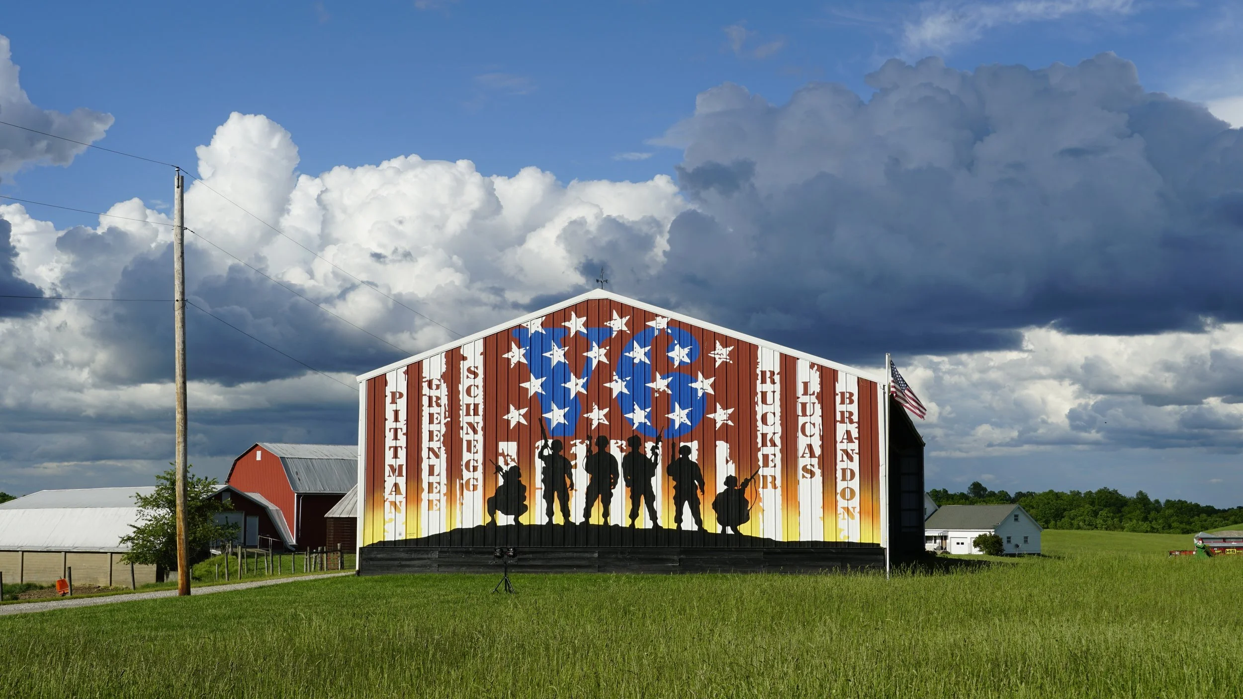 A barn decorated with silhouettes of soldiers and a large American flag design on its front, located in a rural Beallsville Ohio area under a partly cloudy sky.
