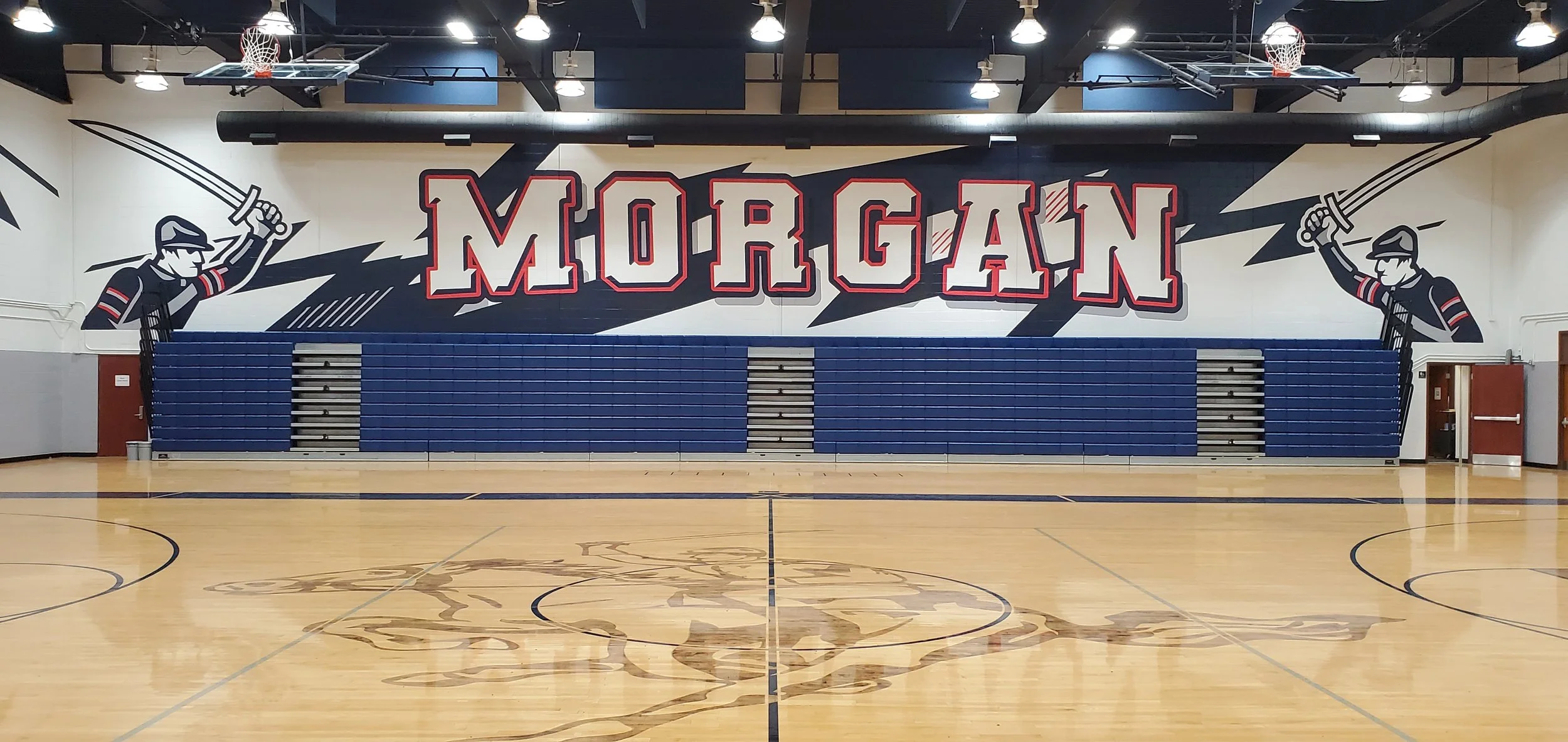 Empty gymnasium with a mural of the word 'MORGANT' in large white and red letters, flanked by illustrations of firefighters holding swords, and blue bleachers against the wall.