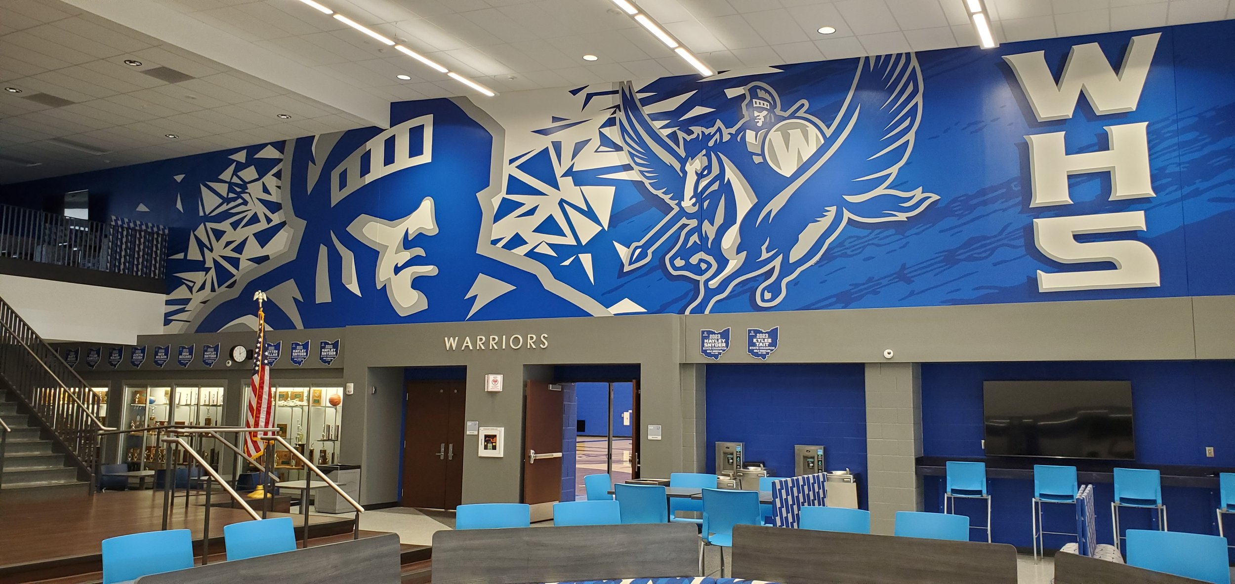 Interior of a sports arena with a large mural on the wall featuring a basketball player, a warrior with wings, and the words "WARRIORS" and "WAS". There are blue chairs, a staircase, and a trophy display case visible.