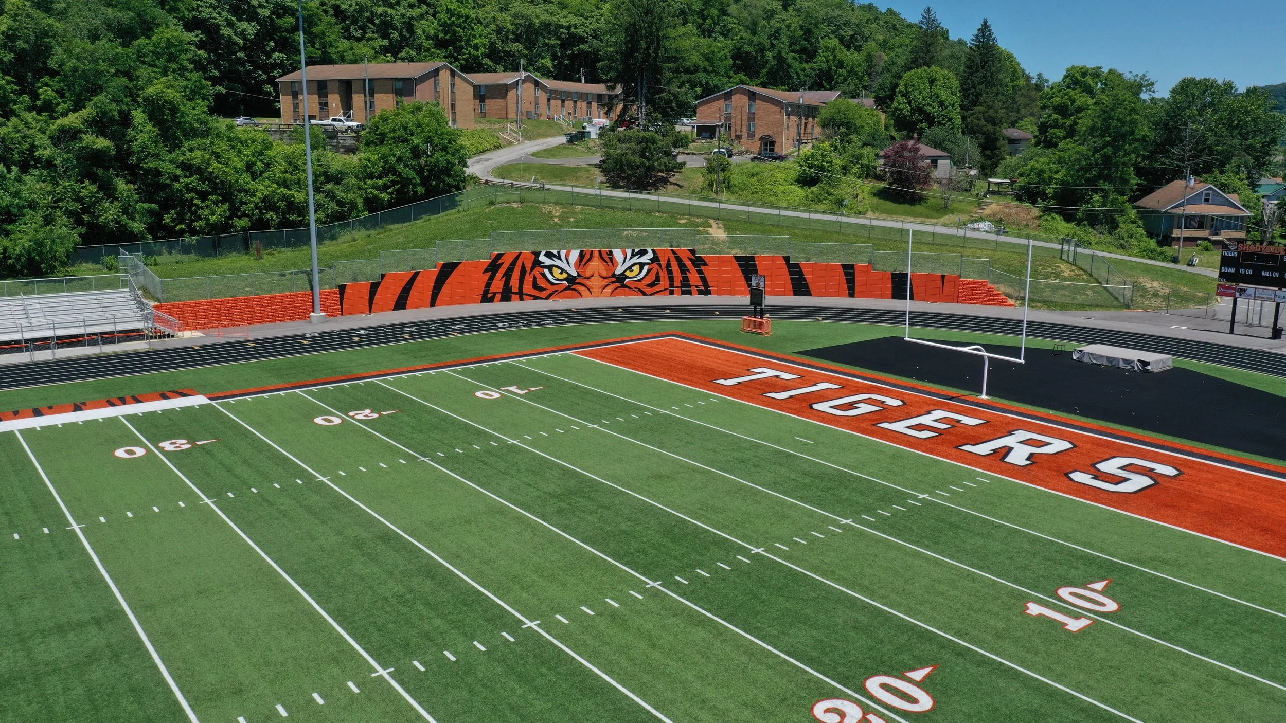An empty football field with artificial turf, featuring a large tiger face mural on the end zone wall. The field has yard lines and numbers marked, with a running track surrounding it. In the background, there are residential buildings and green tree