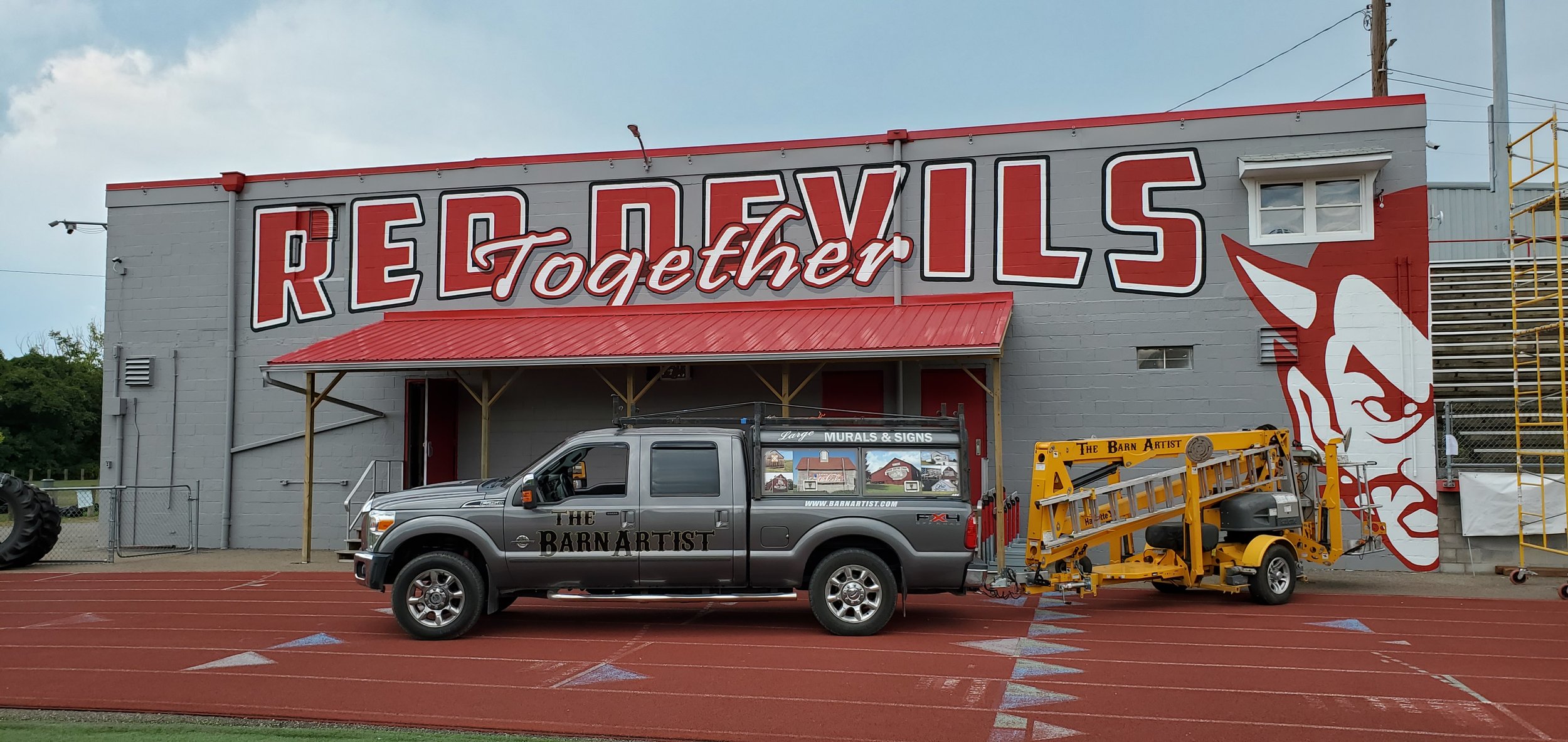 A large building with a mural reading 'RED DEVILS together' in big letters, with a cartoon devil's face on the right side of the wall. In front, there's a gray truck with the words 'The Barn Artist' on the side, parked on a red sports track surface, 