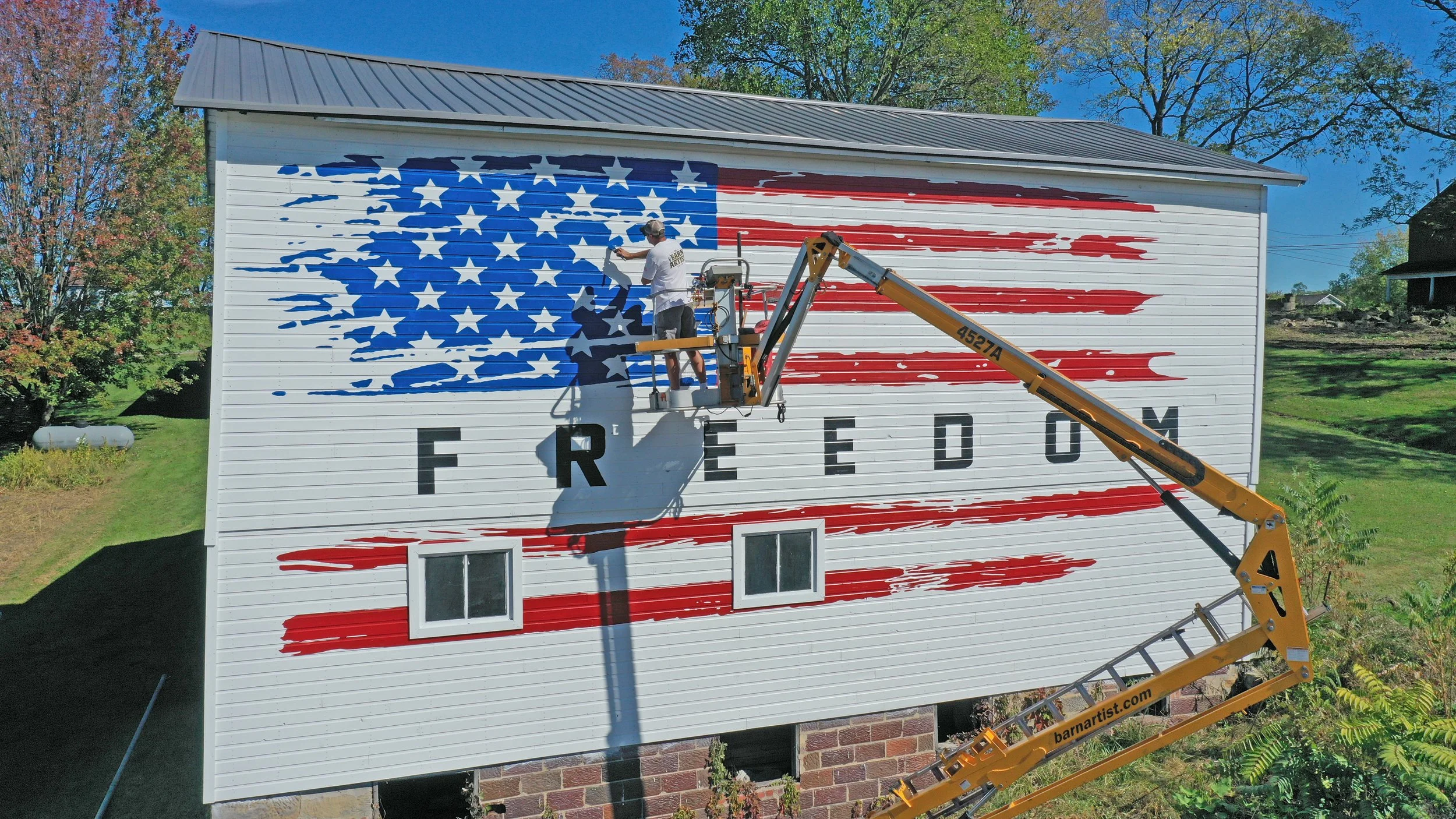 A person painting a mural of the American flag on the side of a white house with the word 'FREEDOM' underneath. The person is standing in a lift with a raised platform, wearing a baseball cap and white T-shirt. The mural features red and white stripe