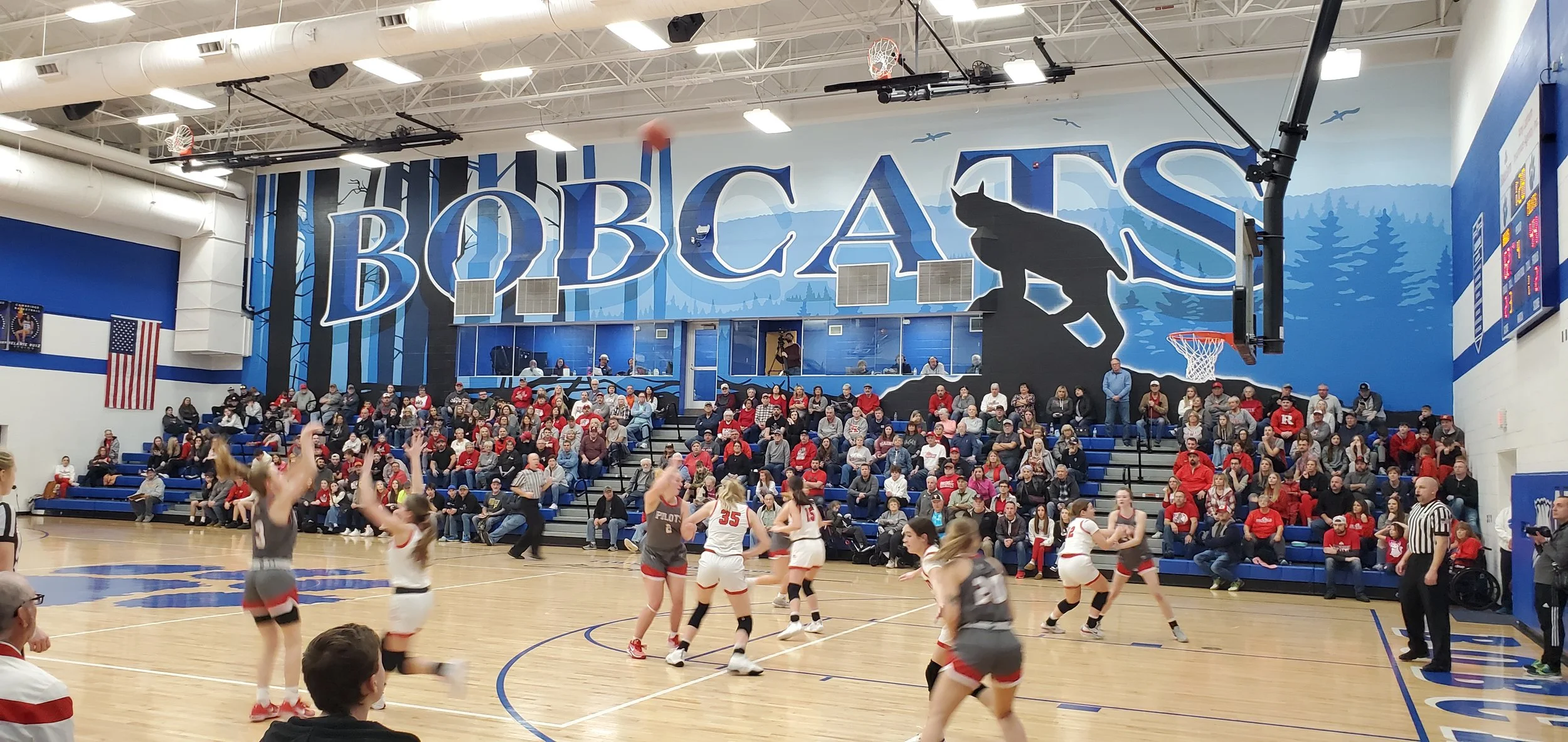 A basketball game taking place in a gymnasium with spectators seated on bleachers. The wall features a large mural with the word 'BOBCATS' and a bobcat silhouette. Players are actively playing on the court, some wearing white and others in gray unifo