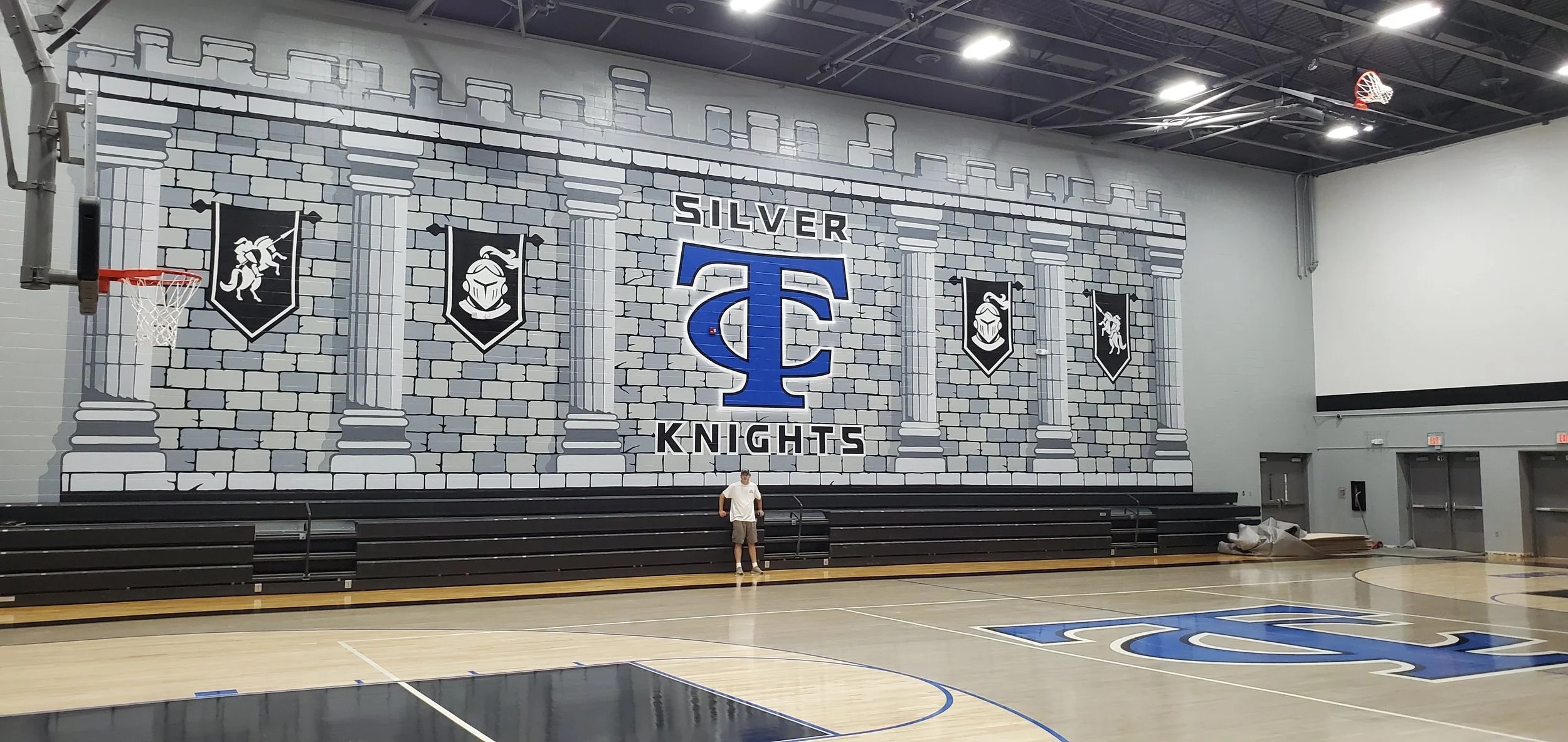 An indoor basketball court with a mural of a castle wall and columns. The mural features the logos of the Silver Knights, including a large blue letter 'T' and the word 'Knights,' as well as shields with images of knights. There is a basketball hoop 