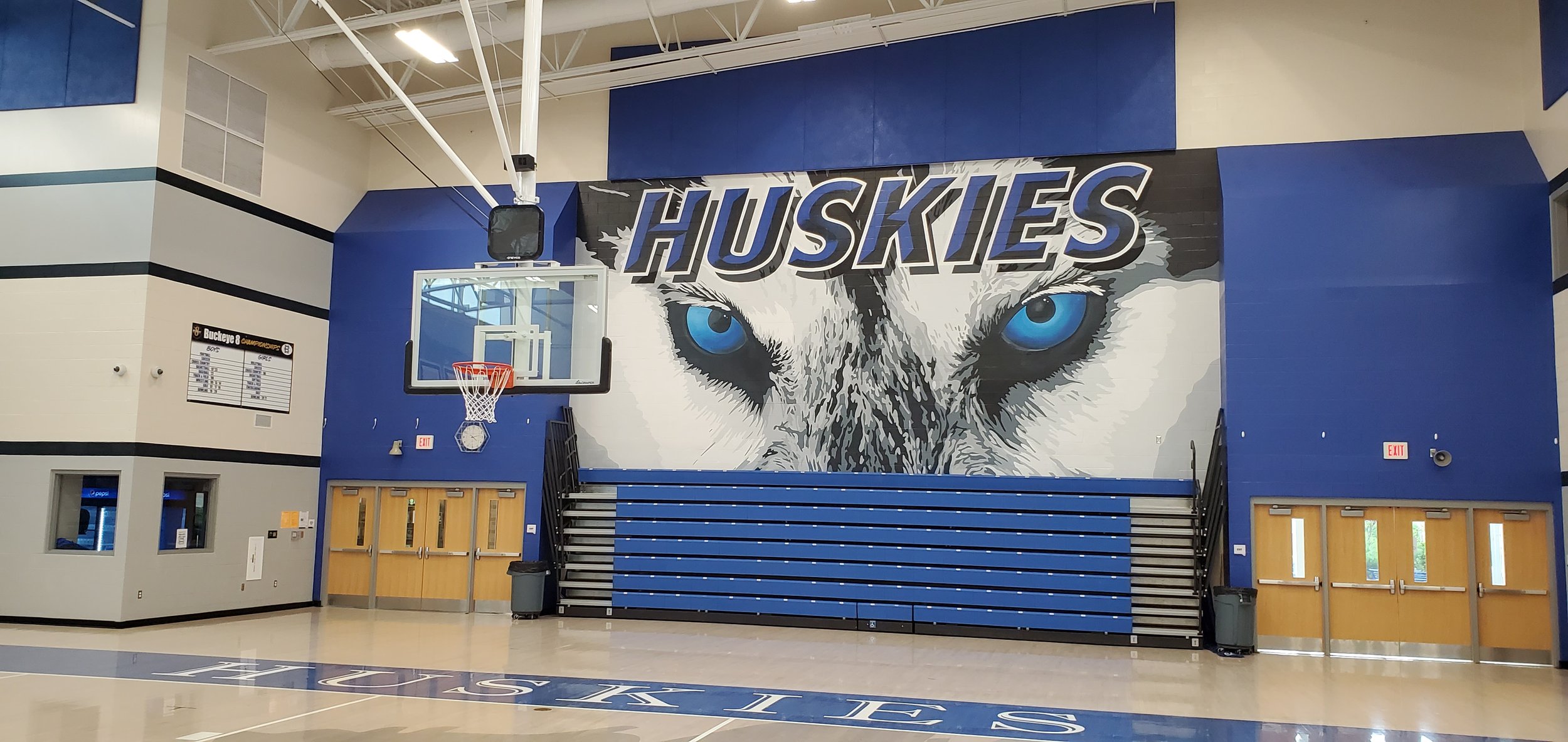 An indoor gymnasium with a large mural of a husky dog's face and the word 'HUSKIES' above it. There is a basketball hoop, blue bleachers, and a blue and white color scheme.