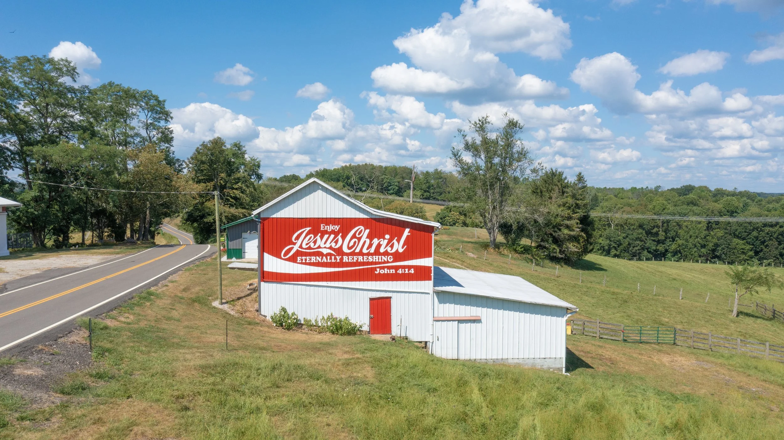 A white barn with a red and white sign that reads "Enjoy Jesus Christ Eternally Refreshing" and references John 4:14, located beside a winding rural road with green trees and rolling hills in the background.