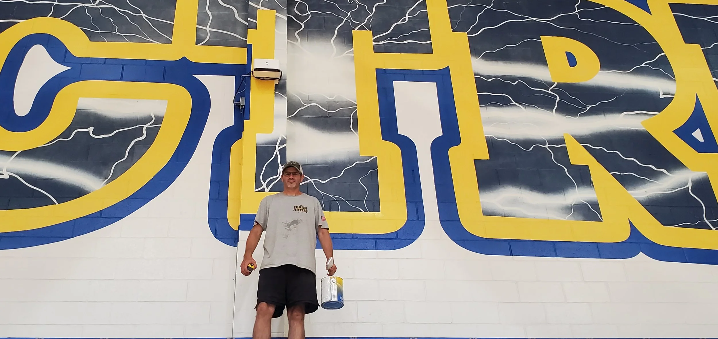 Man standing in front of a large mural with yellow and blue outline letters and lightning storm background. Man wears a gray t-shirt, black shorts, and a cap, holding a paint bucket and a paintbrush.