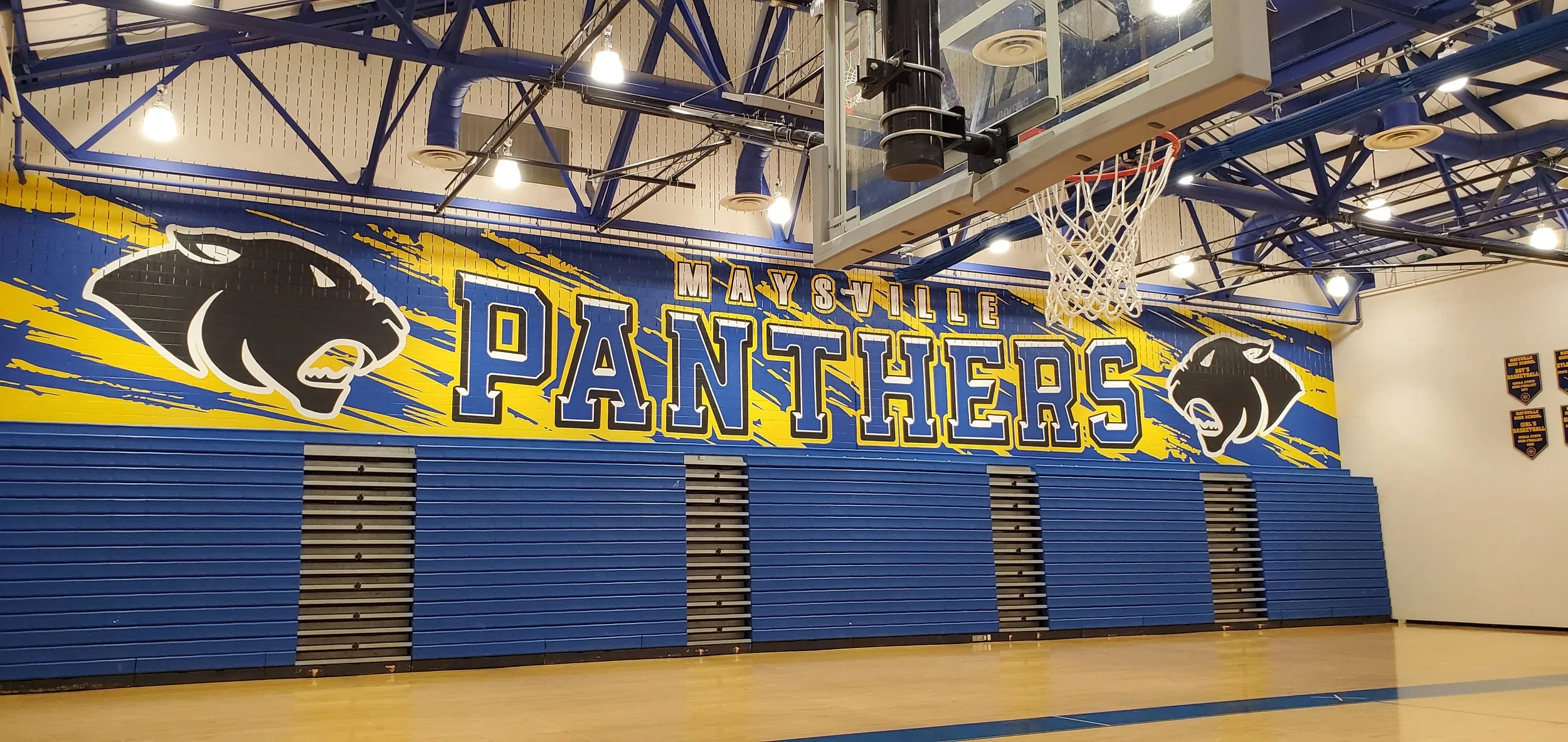 Indoor gymnasium wall with a large sign that reads 'MAYSVILLE PANTHERS' in blue and yellow, featuring two panther logos and a basketball hoop mounted on the wall.