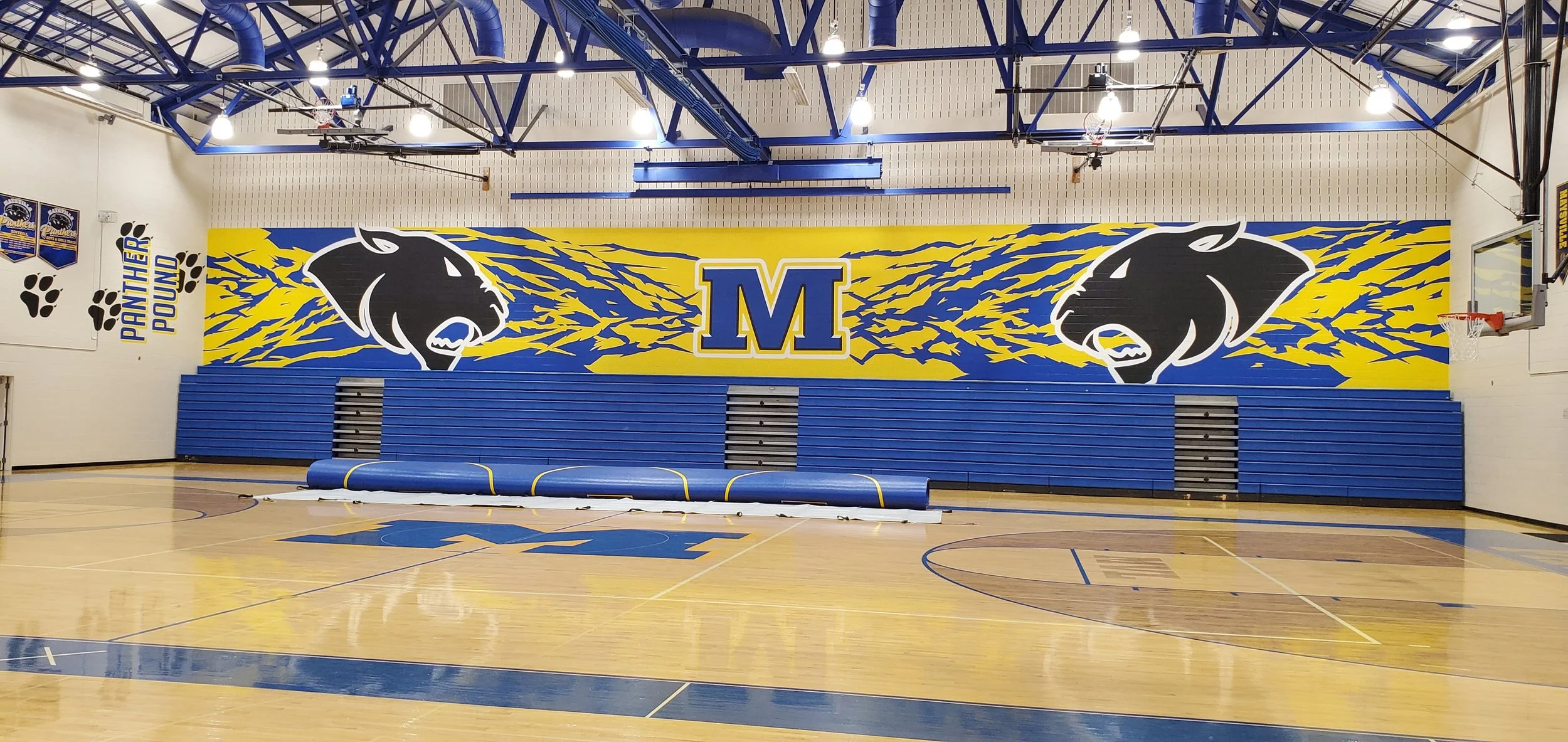 An indoor gymnasium with blue and yellow Panther-themed decorations. The wall features two panther mascots facing each other with a large blue and yellow 'M' in the center. There are blue bleachers folded up against the wall and basketball hoops on e