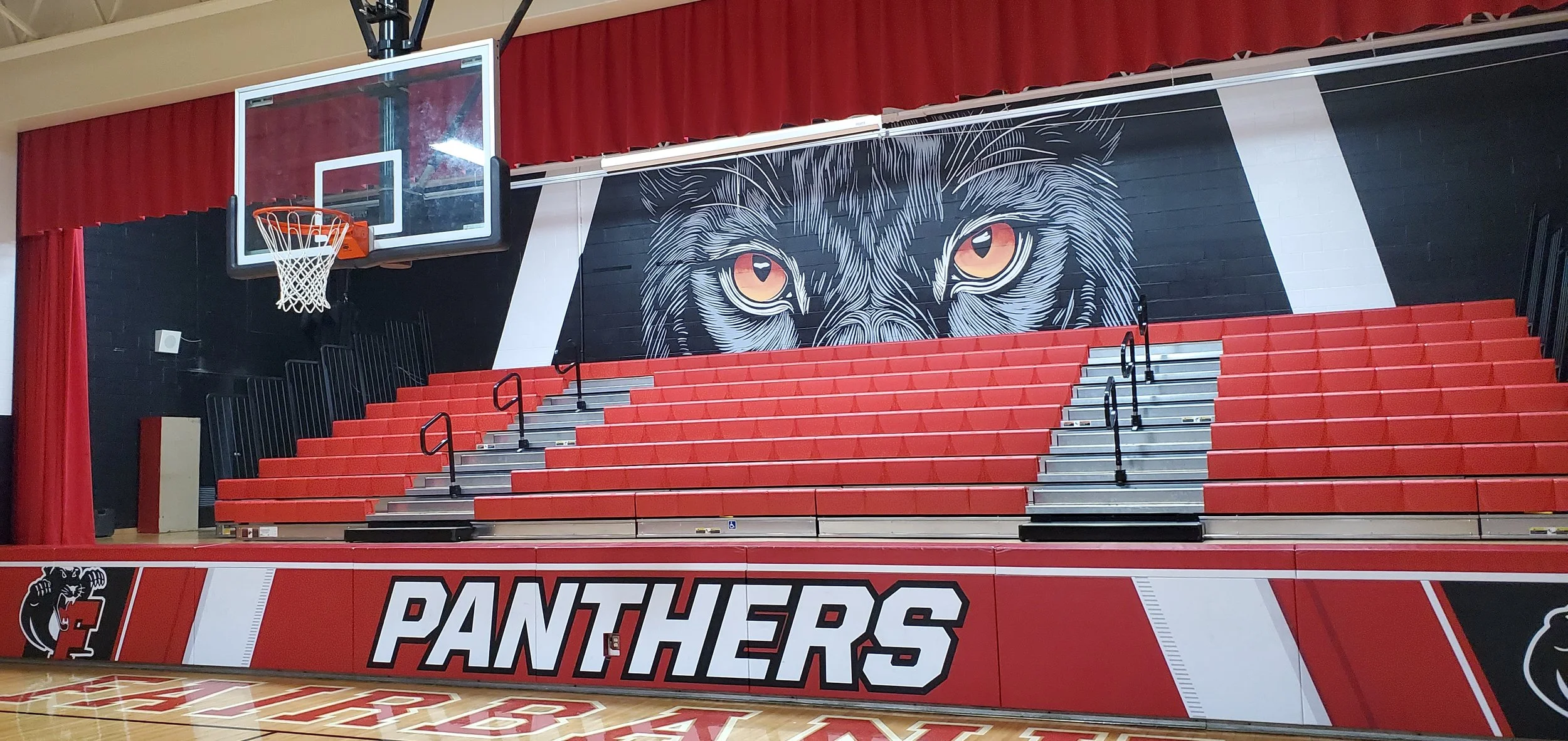 Indoor basketball court with red bleachers, a black wall featuring a large wolf head mural with orange eyes, basketball hoop, with 'PANTHERS' written on the padded wall at the court's edge.