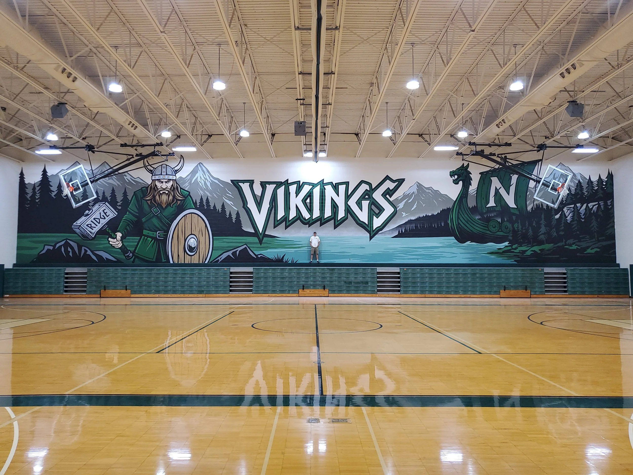Indoor basketball court with painted murals of Vikings, mountains, and trees on the wall. One person standing in front of the mural. Overhead lights on the ceiling.