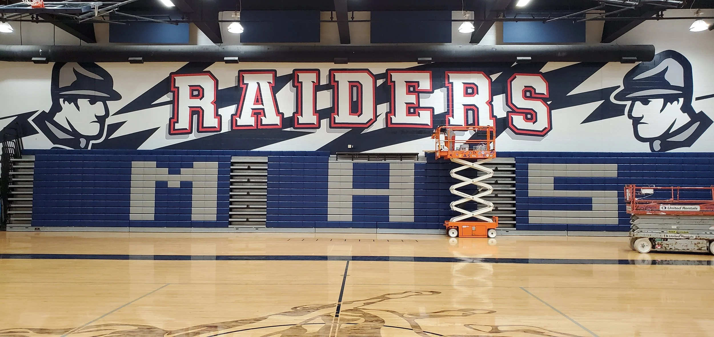 A school gymnasium wall decorated with a large sports-themed mural of the Raiders team logo, including light blue and dark blue seats and a lift platform in front of the wall.