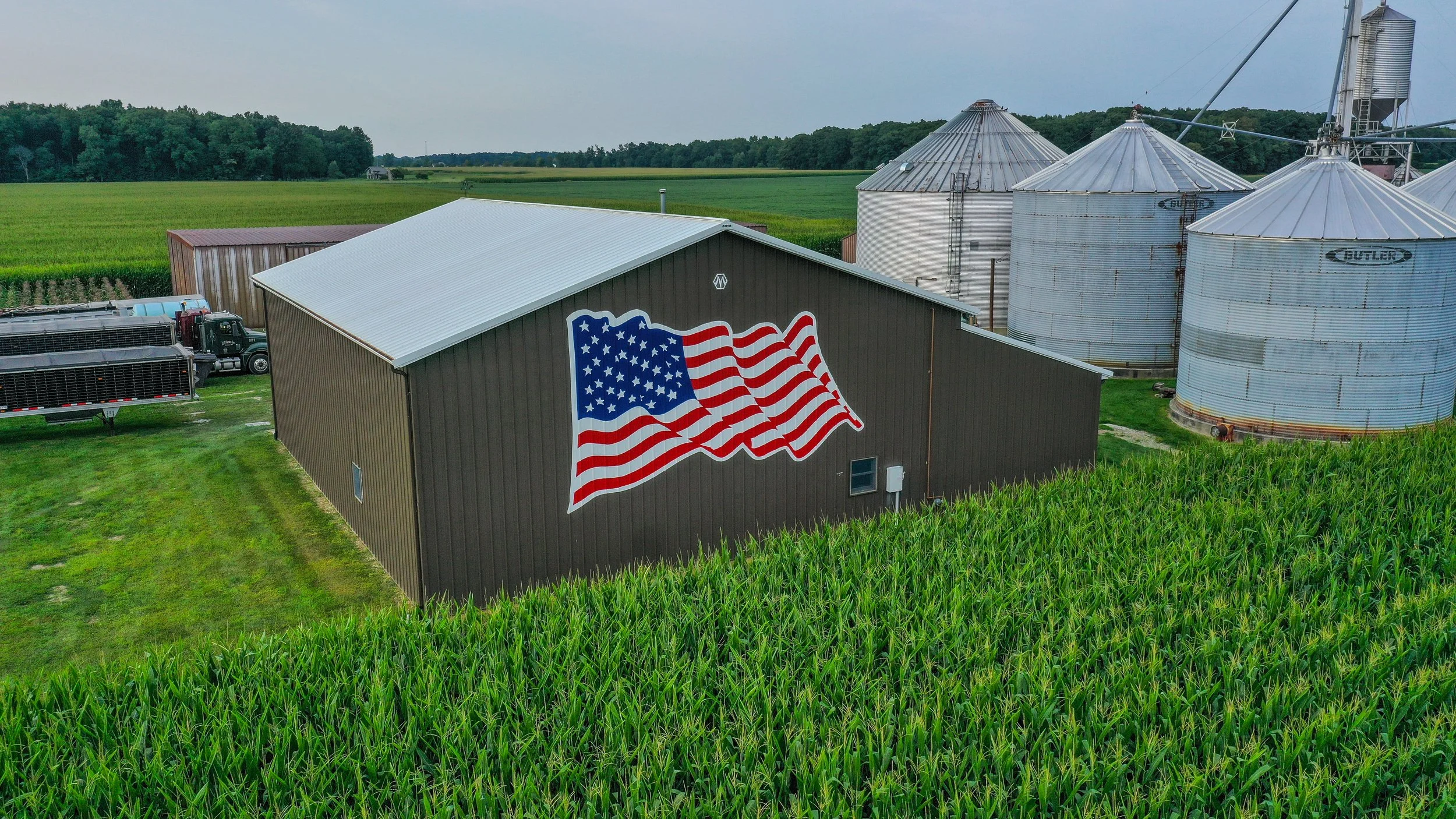 A farm with a barn displaying a large American flag on its side. There are silos and farm equipment nearby, and green fields extend into the distance.