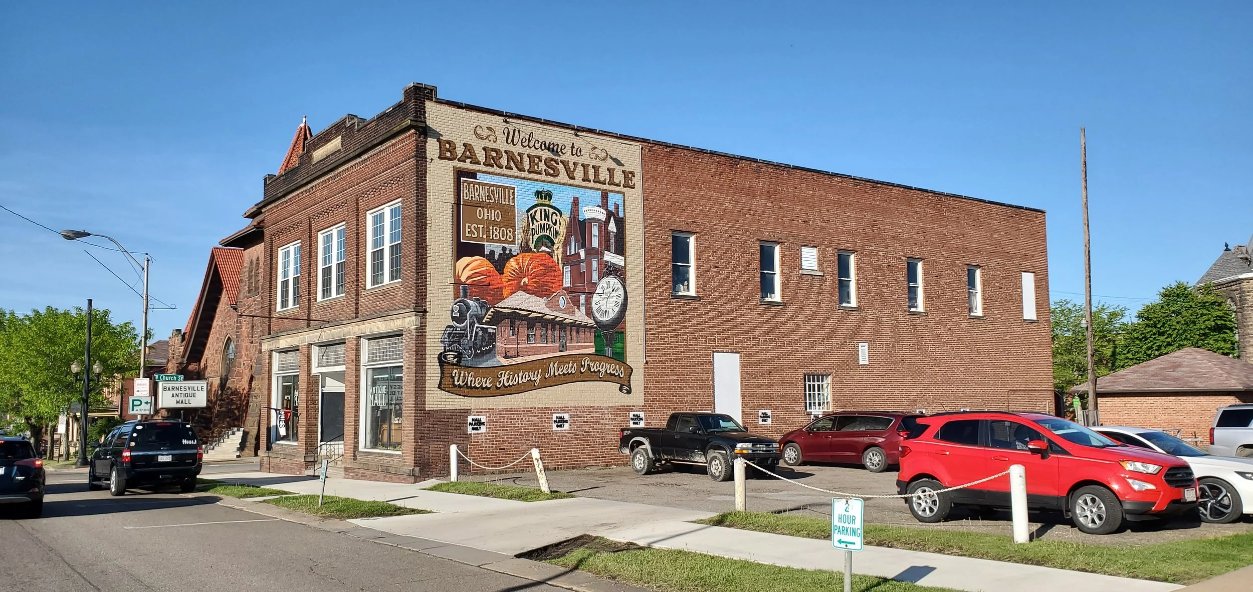 Brick building with mural welcoming visitors to Barnesville, Ohio, established 1808, with historical and pumpkin imagery, parked cars in front, green trees, street signs, and clear blue sky.