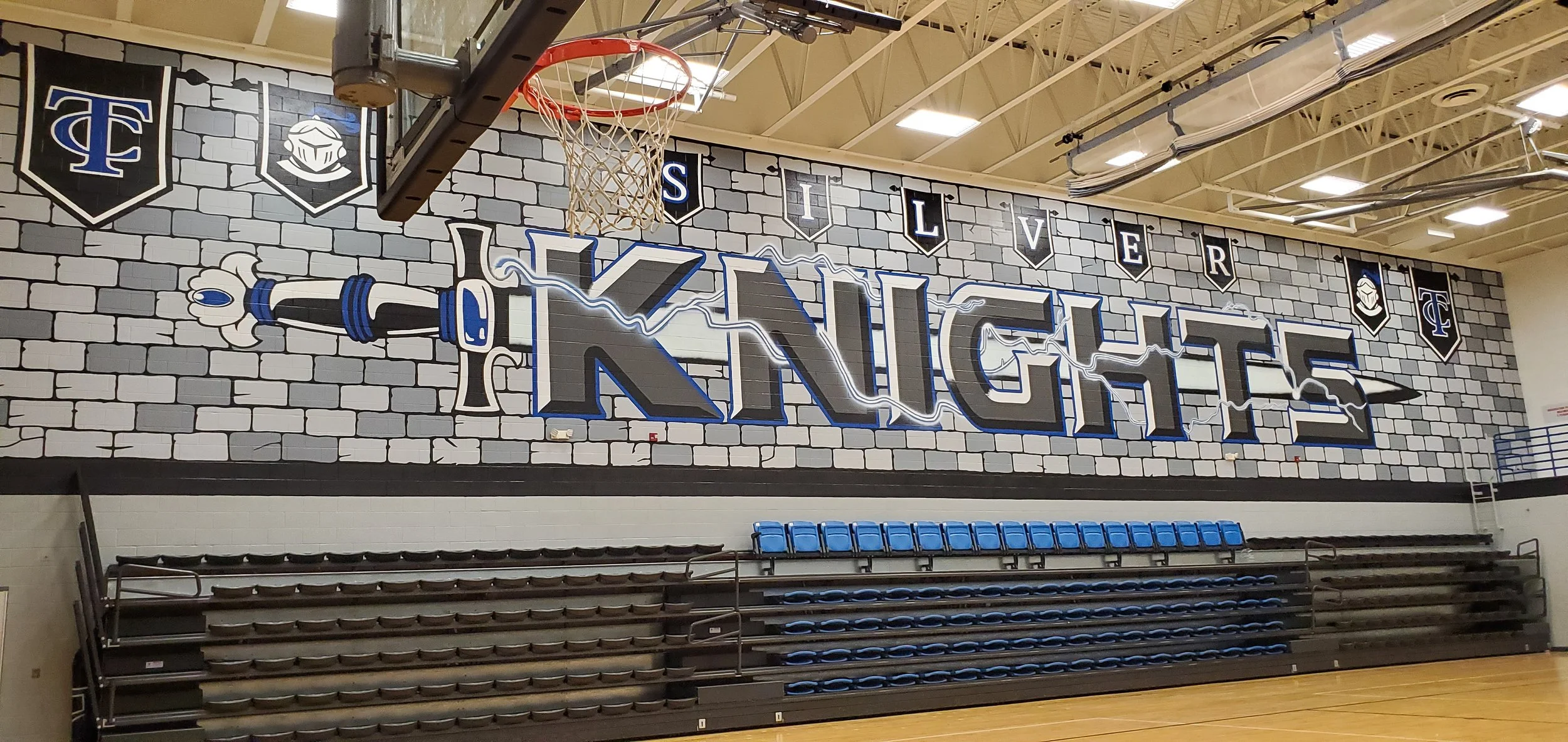 An indoor basketball gym with a wall mural that reads 'Knights' featuring a large sword, lightning bolts, and the words 'Live' and 'Love' on banners. There are blue bleacher seats and basketball hoops.