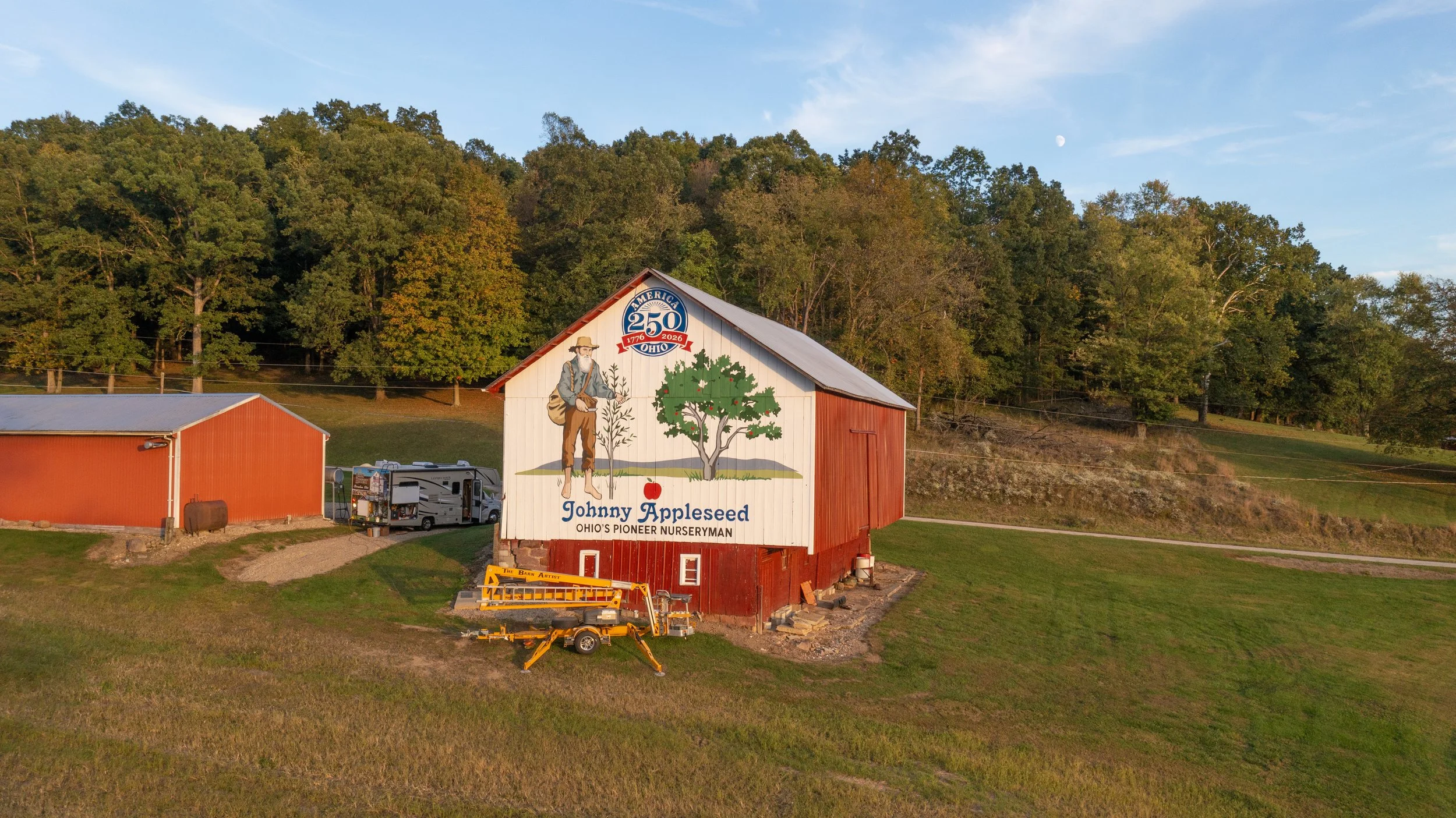 A red barn with a mural depicting a man named Johnny Appleseed, celebrating Ohio's pioneer nurseryman, with trees, an apple, and a 250th anniversary sign in the background.