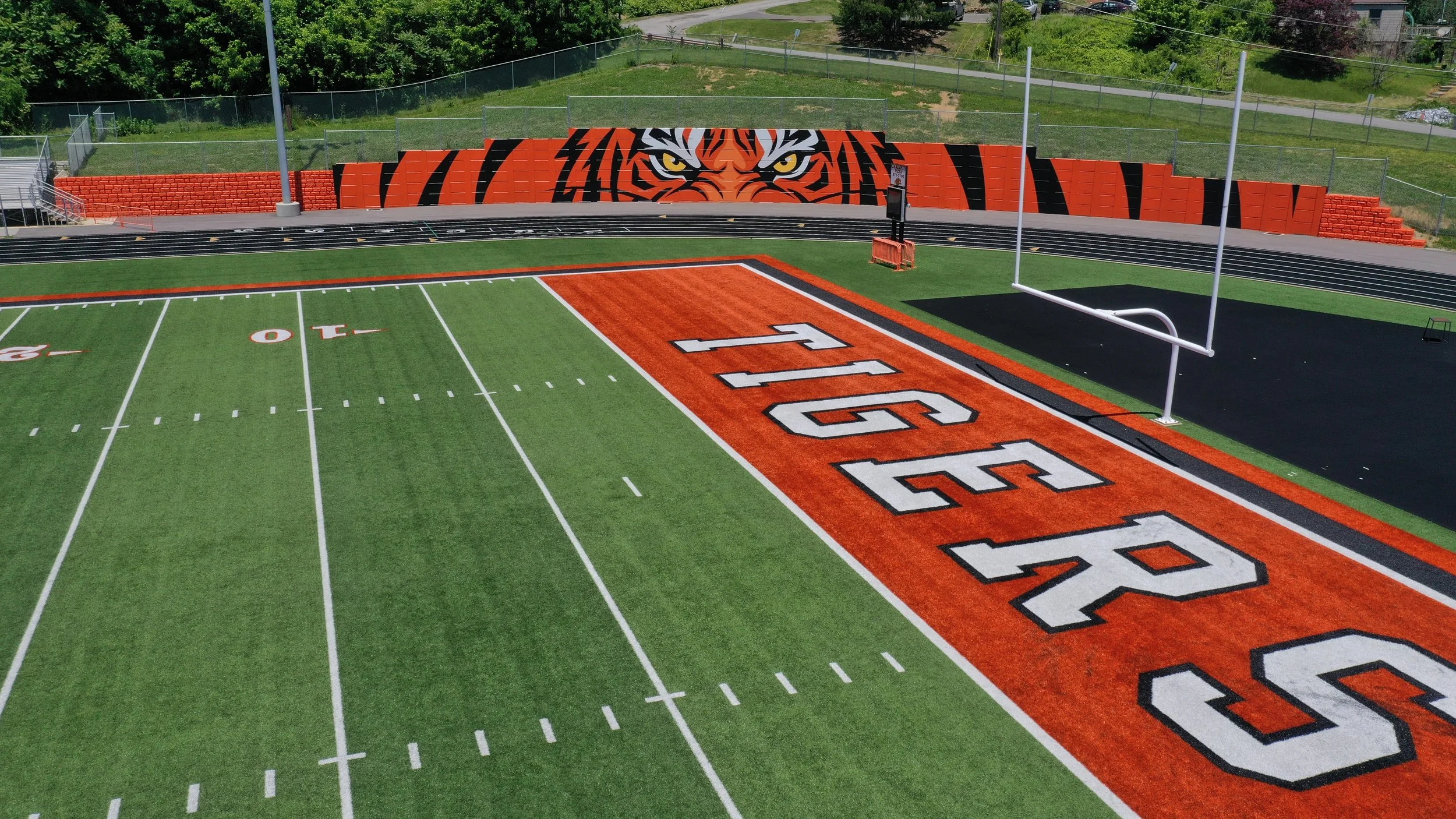 An empty athletic field with a running track and a football field featuring a large tiger mascot painted on the wall behind the goalposts, with green trees and grass surrounding the area.