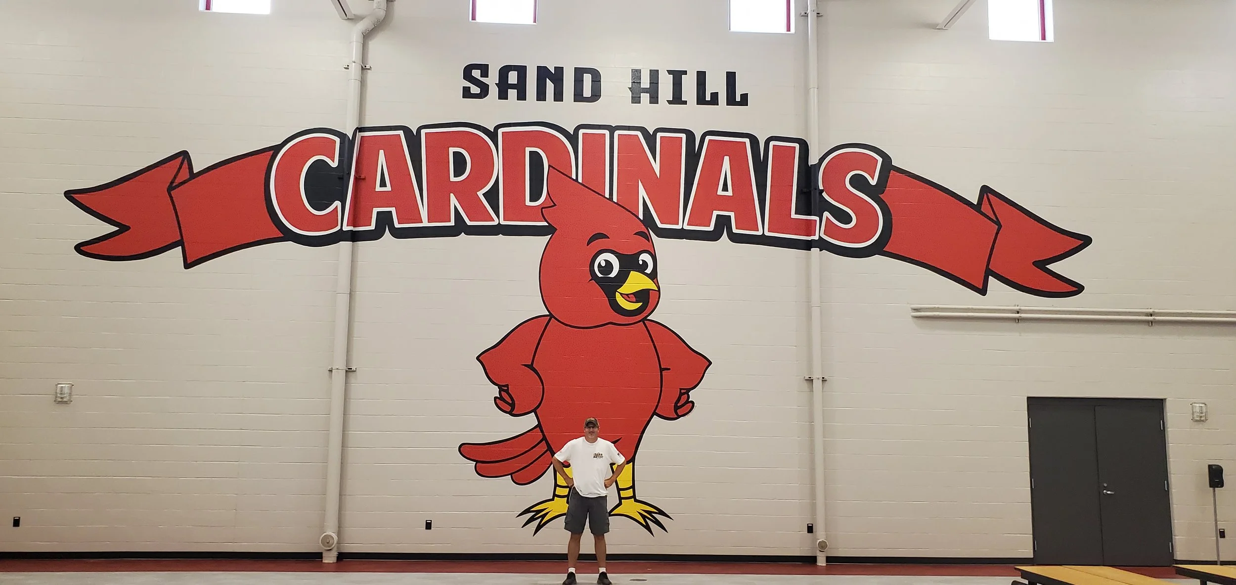 A person standing in front of a large cartoon mascot of a cardinal in the gymnasium at Sand Hill Cardinal school.