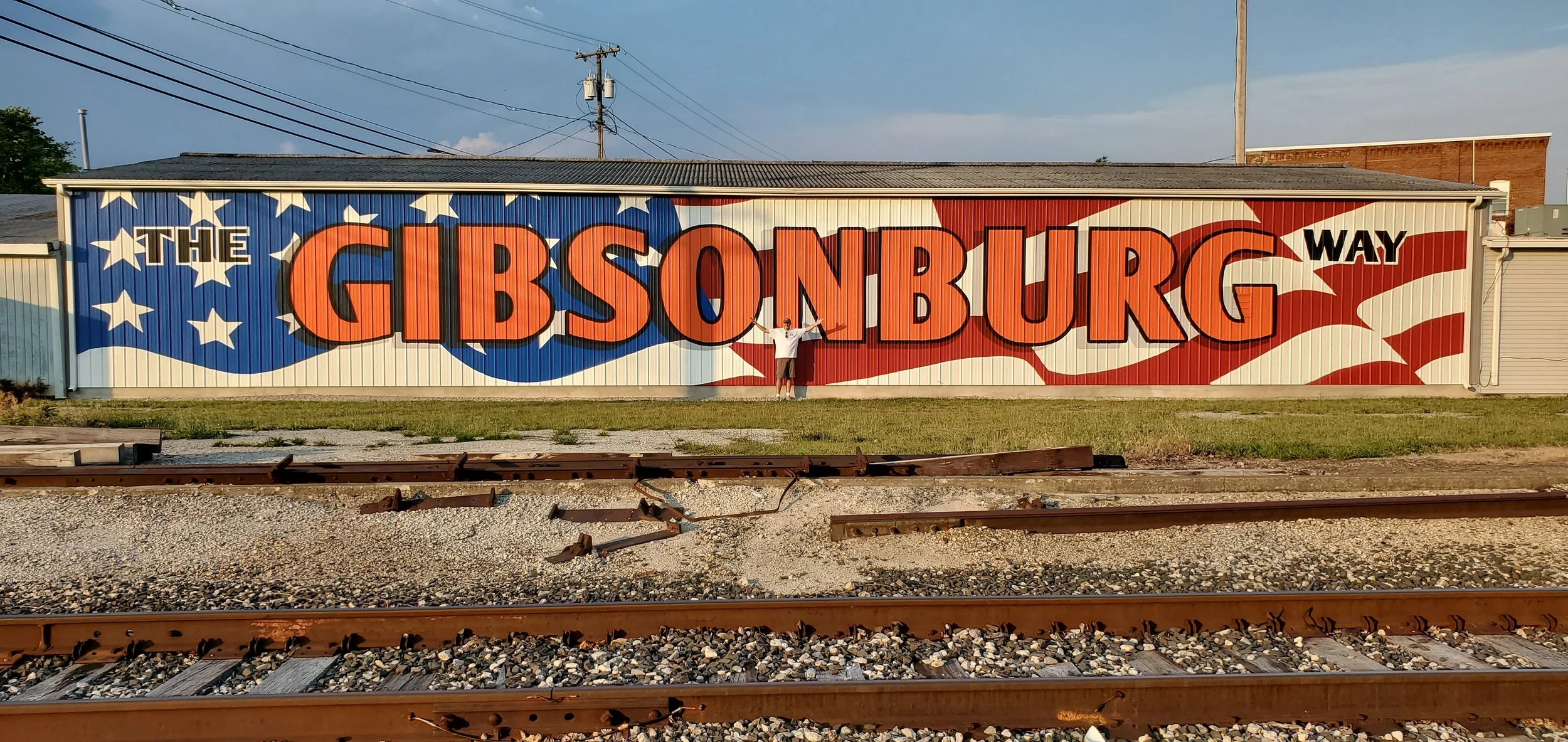 A large mural on a metal building wall with the text 'The Gibson Way' against a patriotic American flag background. A person stands in front of the mural, holding a flag.