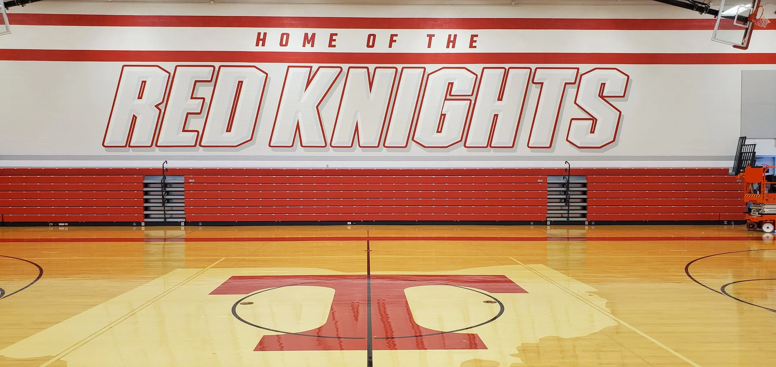 An empty indoor basketball court with red bleachers and a large wall display reading "Home of the Redknights". Basketball hoop is visible in the upper right corner.