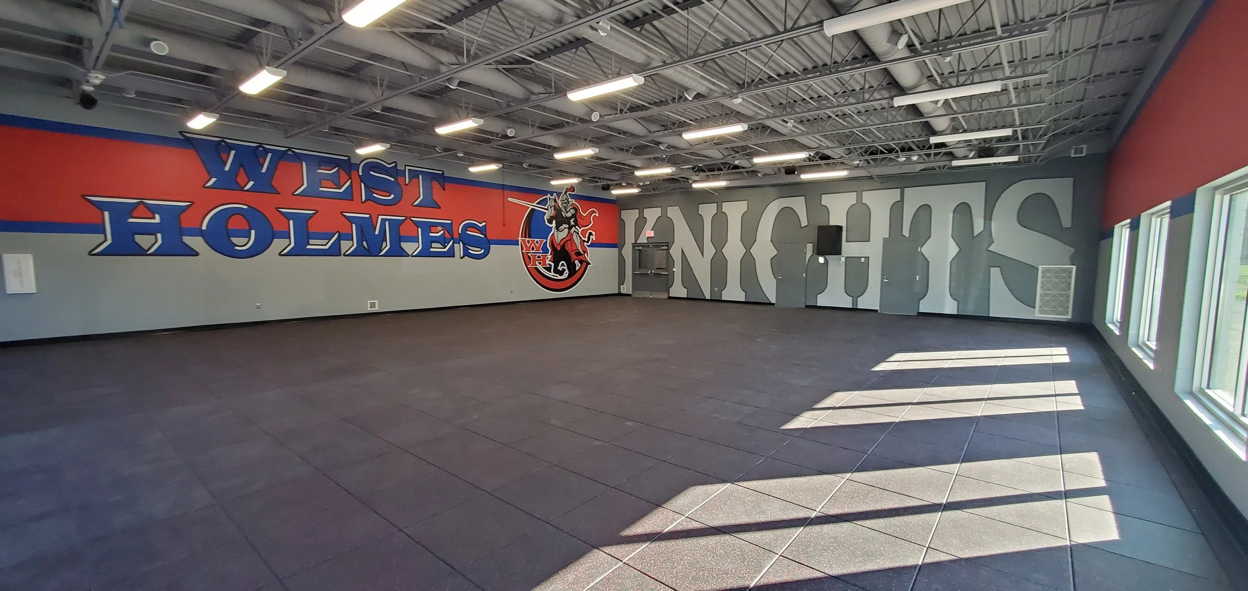 An empty room with a sports-themed mural on the wall showing the words "West Holmes" and a logo of a knight with a sword. The room has gray tiles, large windows, and a ceiling with exposed beams and lights.