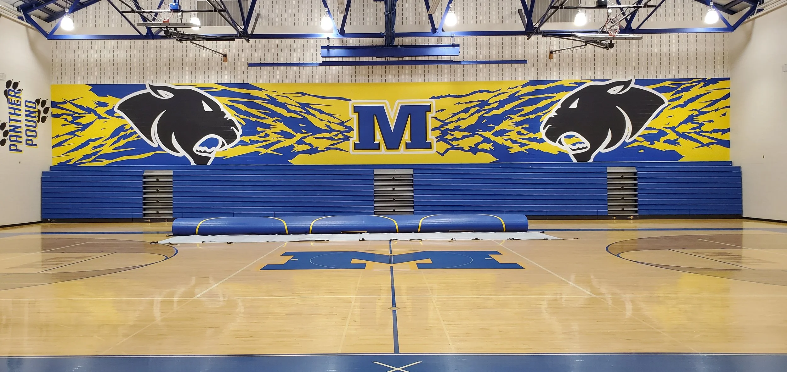 Indoor gymnasium with a large mural on the back wall featuring a black panther head and the letter 'M' in the center, with blue and yellow colors. The gym has blue bleachers, a wooden floor with sports markings, and a rolled-up mat in the foreground.