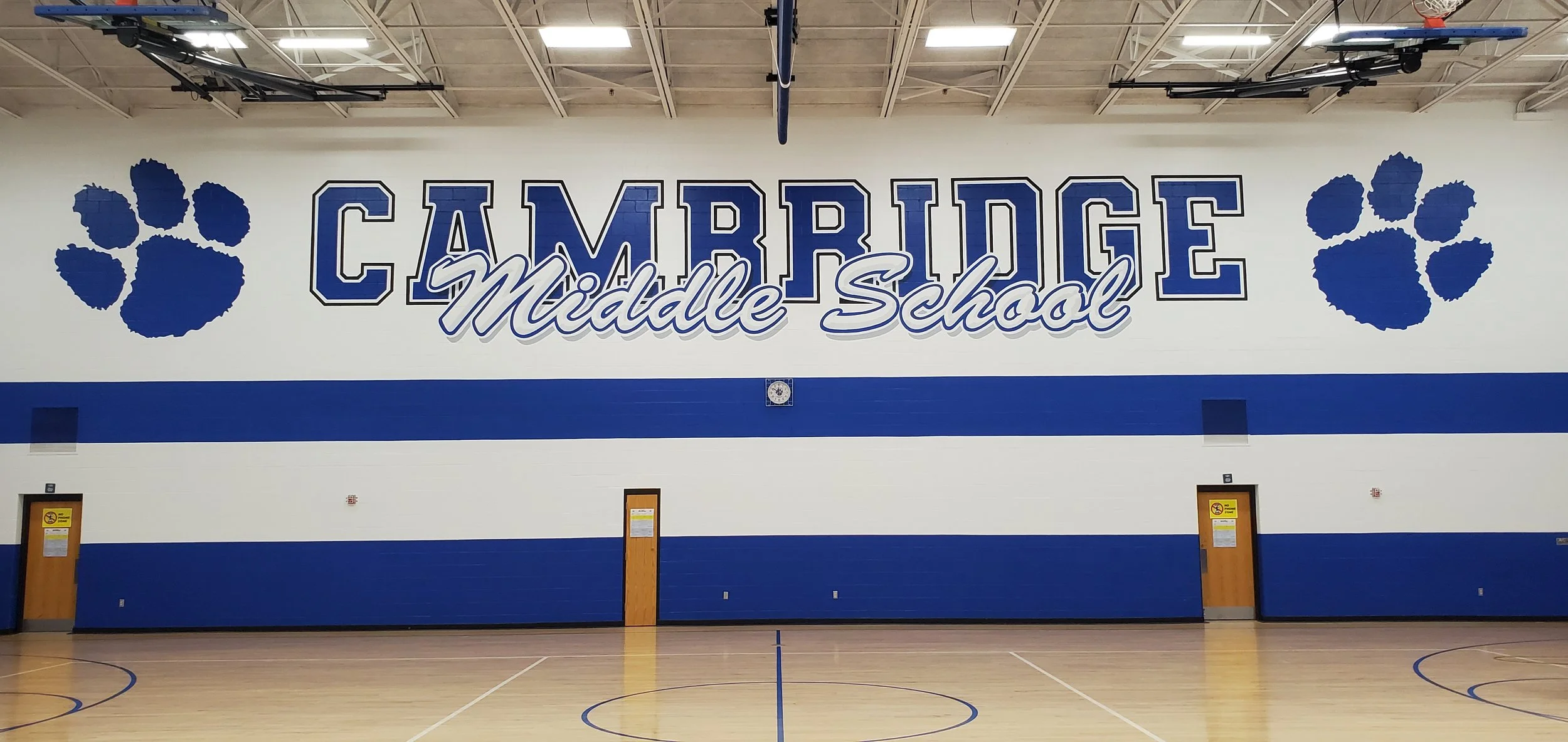 Interior of a gymnasium with a wall sign reading 'CAMBRIDGE Middle School' flanked by blue paw prints; gym floor visible in foreground.