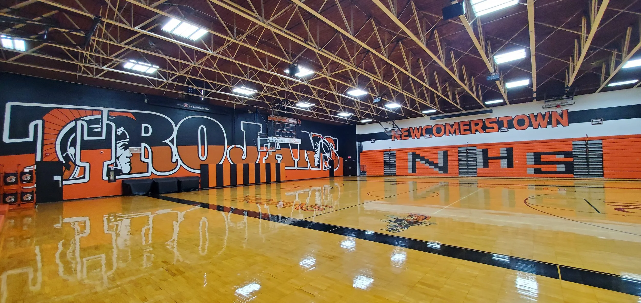 Indoor basketball court with polished wooden flooring, black and orange wall murals, and large orange lettering spelling "NEWCOMERSTOWN."