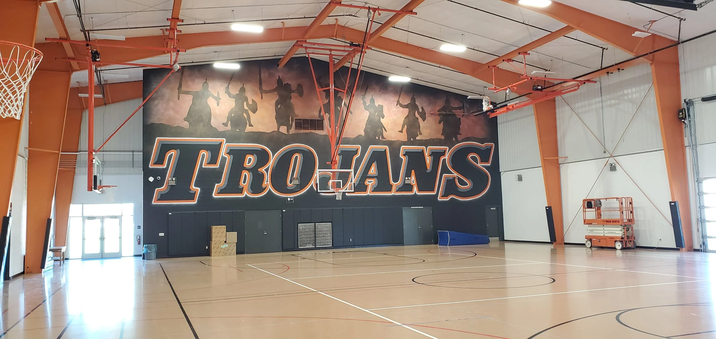 Indoor basketball gymnasium with a large wall mural of five warriors holding swords and shields, titled 'TROJANS' in bold letters.