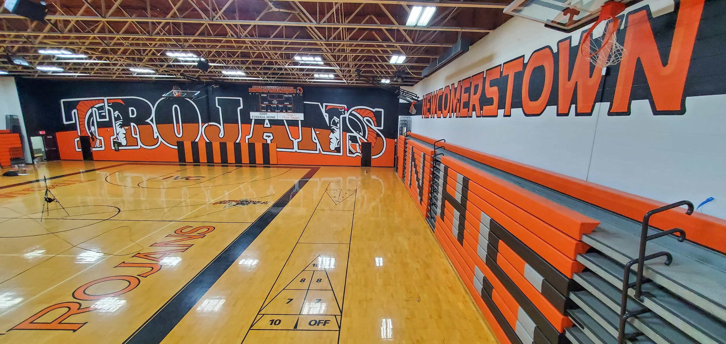 An indoor basketball court with orange and black bleachers on the right side, a large mural in the background featuring a Native American chief with the words 'CROWFIGHTERS' and 'NEWCOMERS' painted on the walls, a basketball hoop with a net, and a sc