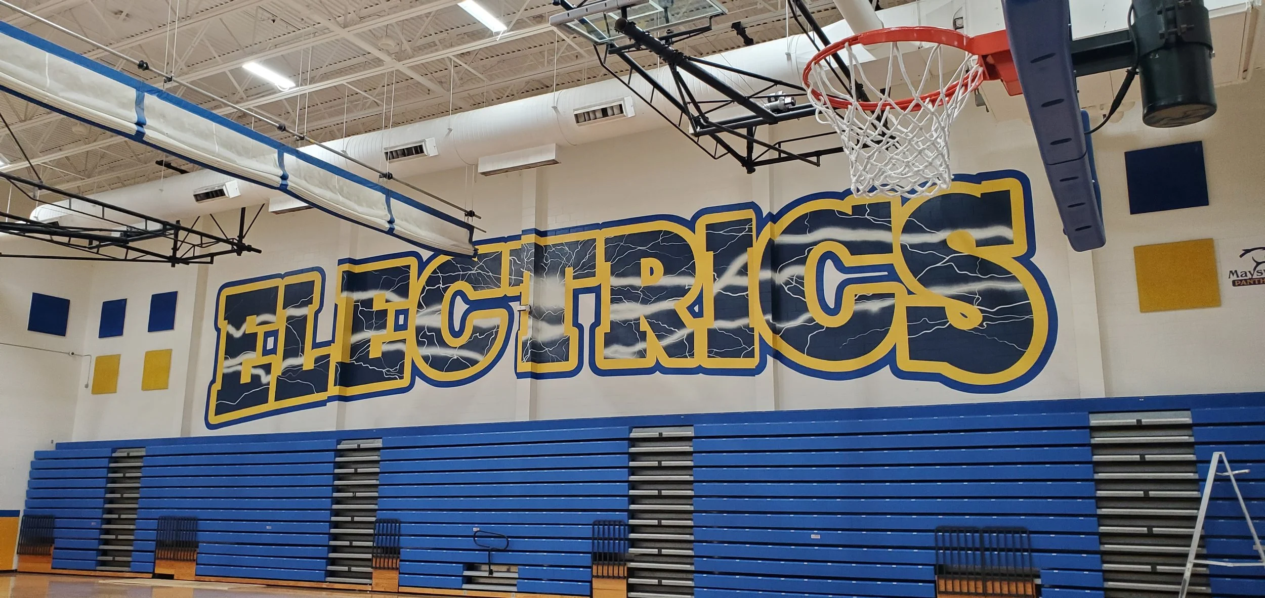 Indoor basketball court with a large wall mural of the word 'Eagles' in bold yellow and blue letters with lightning graphics. There is a basketball hoop with a white net hanging from the ceiling, and blue bleacher seating along the wall.