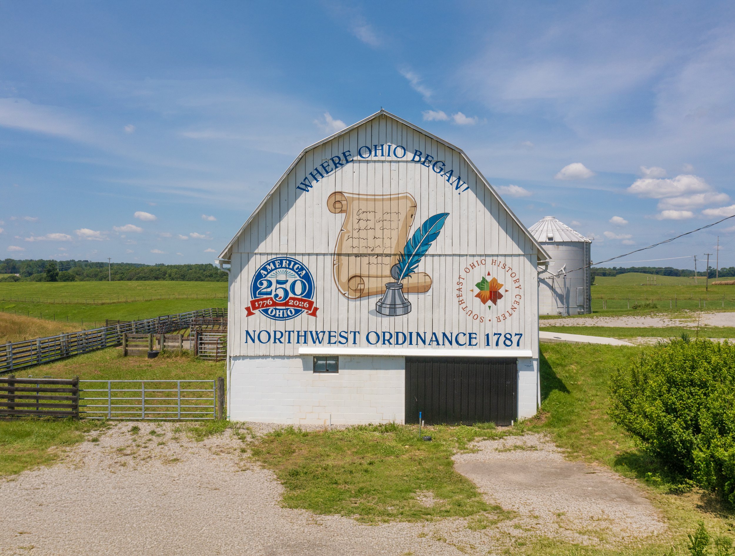 A white barn with a curved roof features colorful signs celebrating Ohio's history and culture, including a 250th anniversary emblem and a quote about Northwest Ordinance, set against a rural landscape with green fields and a blue sky.