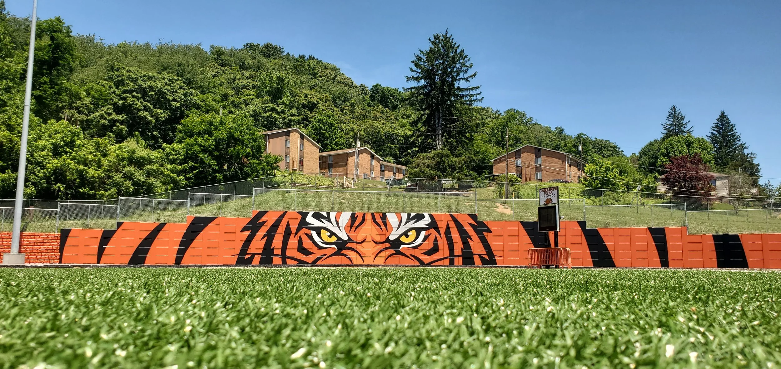 A sports field with a tiger face mural painted on a red and black wall. In the background, there are residential buildings on a hill, surrounded by green trees, under a clear blue sky.