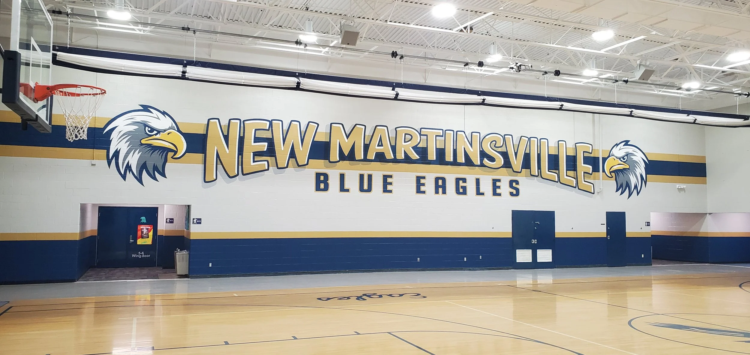 An indoor basketball court with a large sign on the wall reading 'New Martinsville Blue Eagles' accompanied by eagle logos.