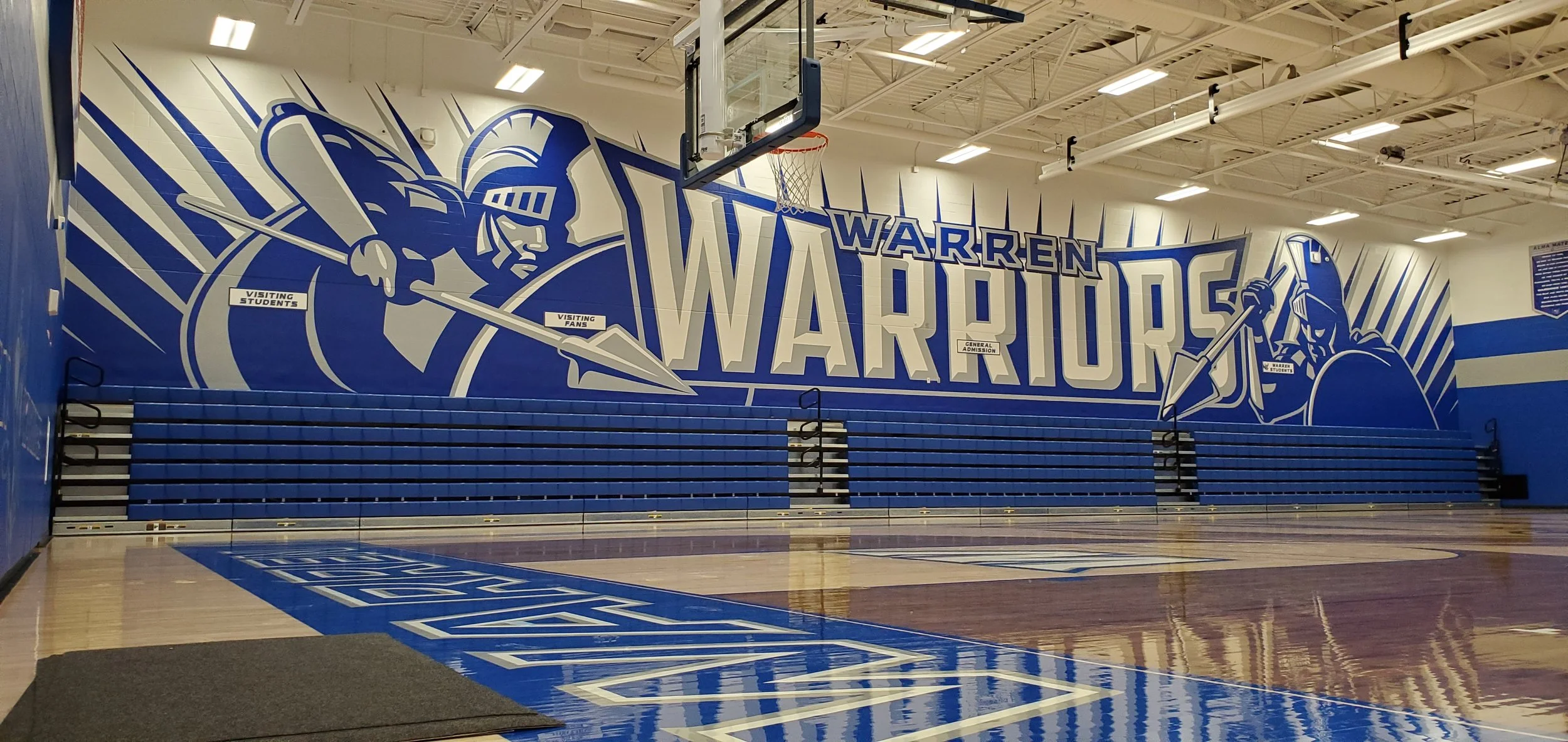 Indoor basketball court with a large blue and white mural of the Warren Warriors team logo on the wall, featuring an eagle and a warrior with a spear. Bleachers are visible in front of the mural, and a basketball hoop hangs from the ceiling.