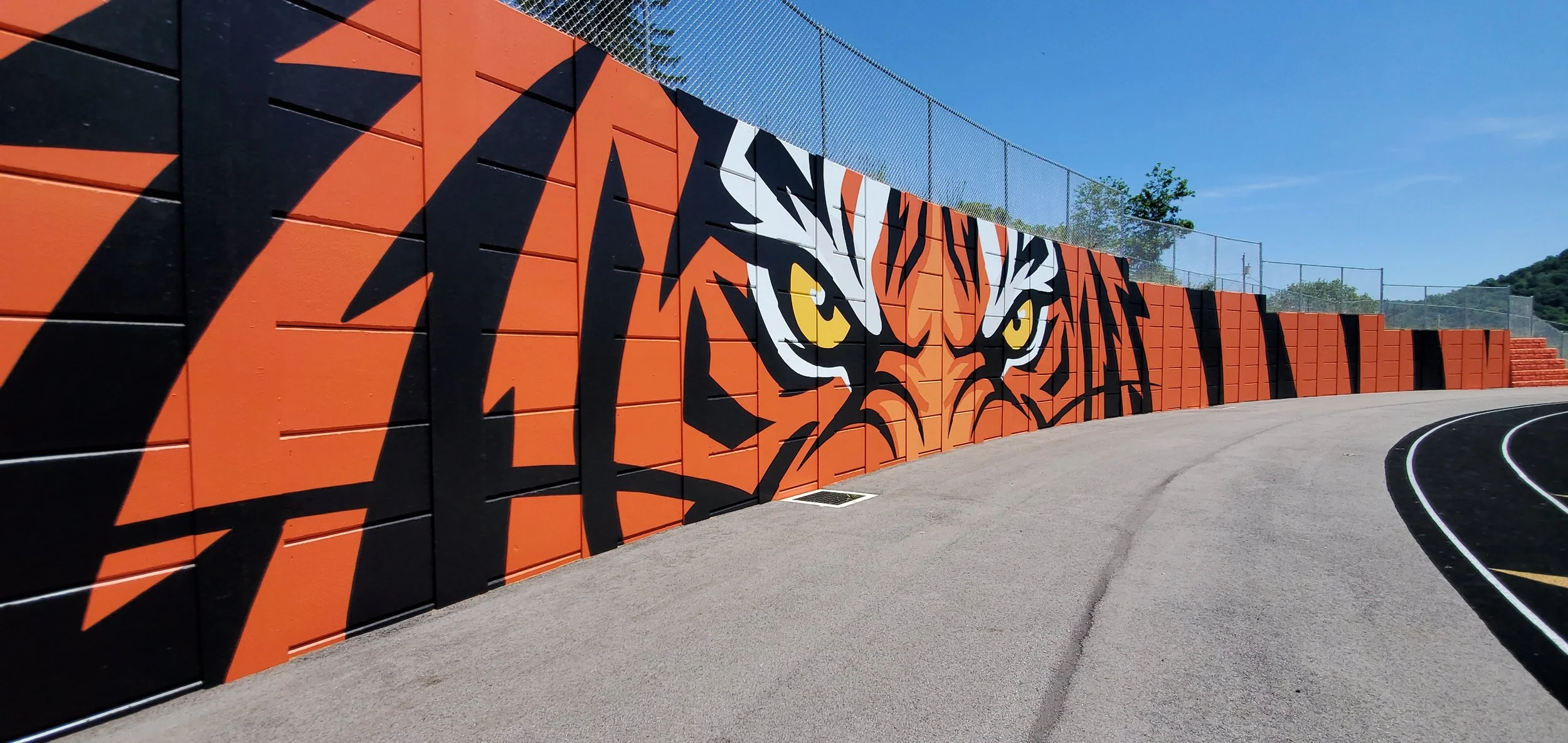 A wall with orange and black tiger stripe mural at an outdoor running track, with a clear blue sky and some trees in the background.