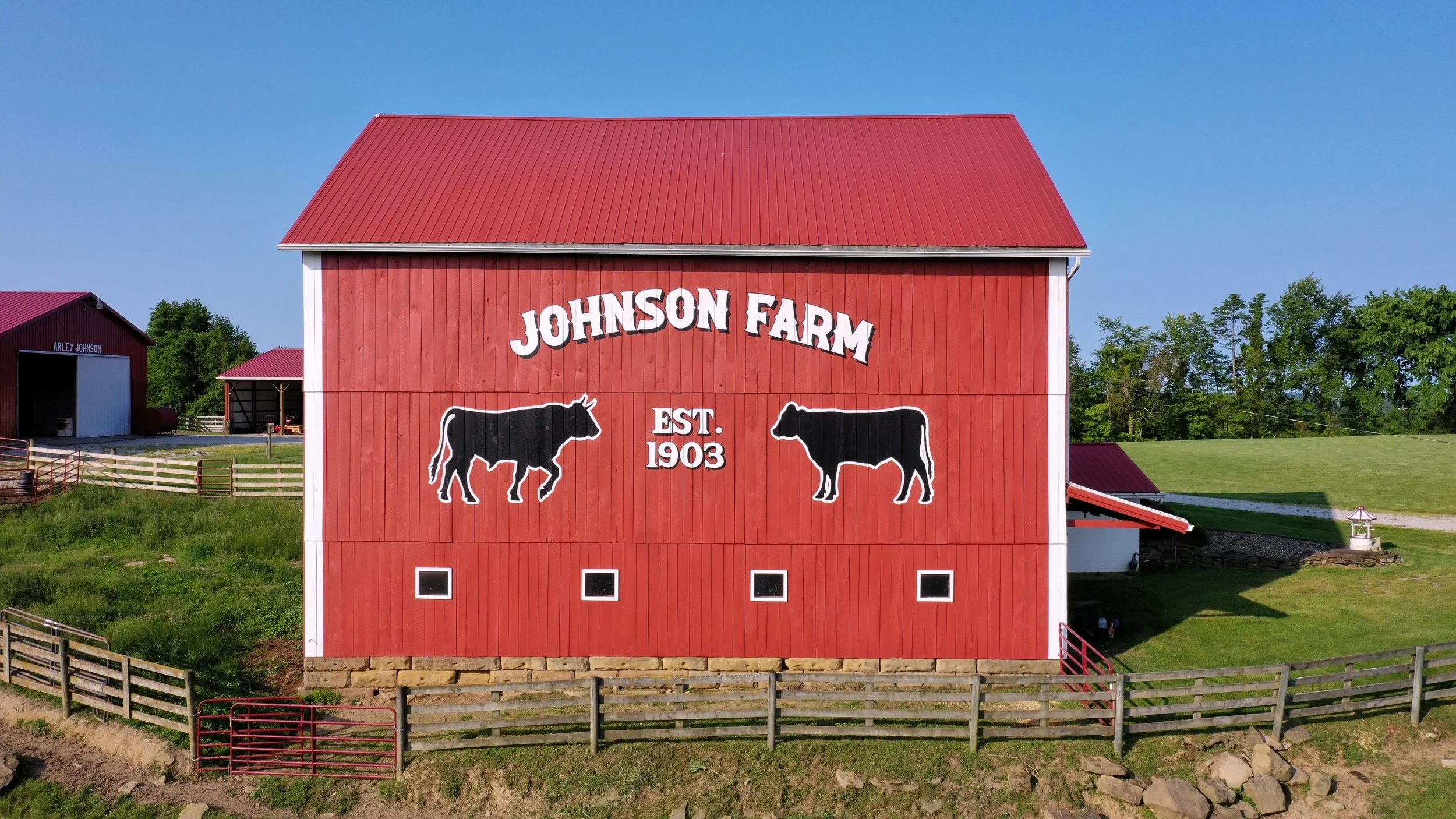 Red barn with white lettering that reads 'Johnson Farm' and 'Est. 1903,' with illustrations of a cow and a calf on the front, set in a rural farm landscape.
