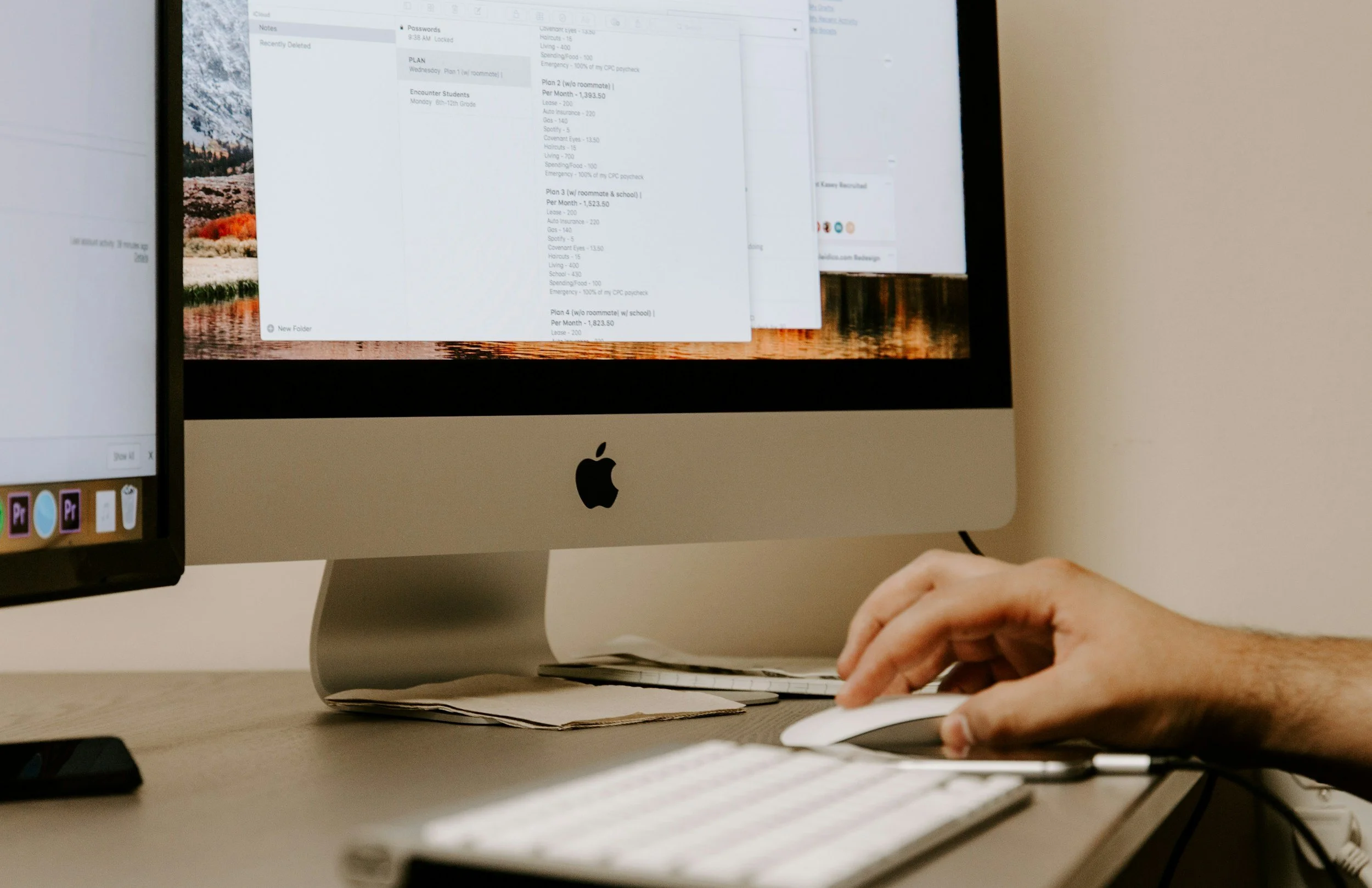 Close-up of a person's hand using a computer mouse in front of an Apple iMac computer displaying a document or spreadsheet on the screen.